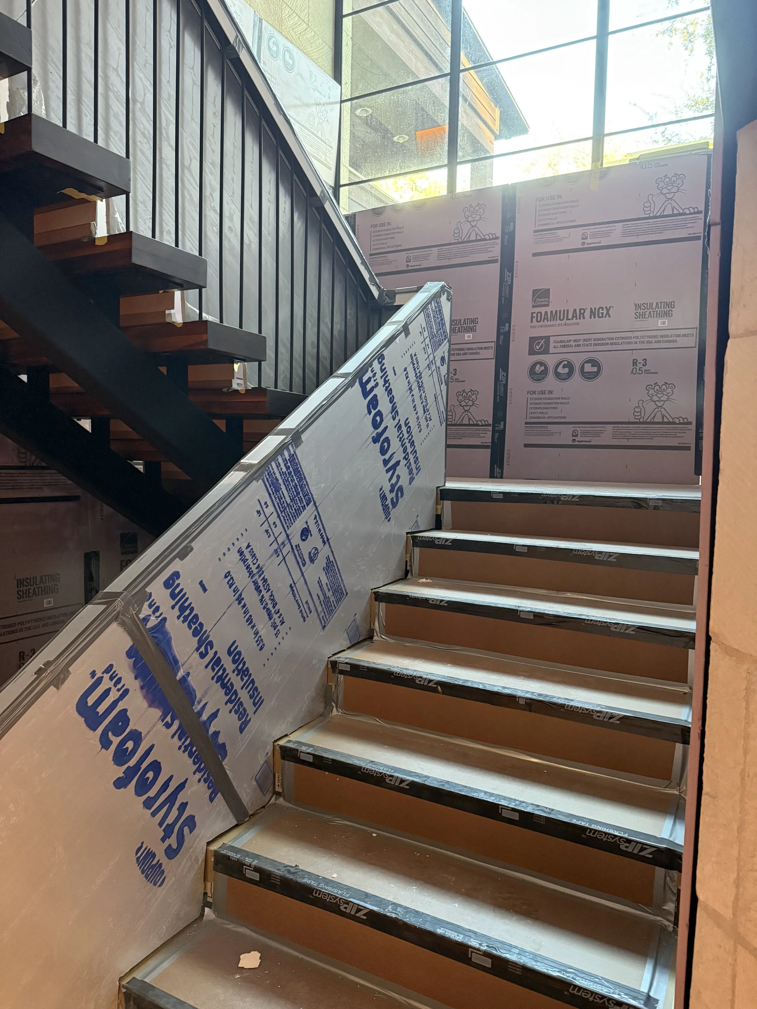 Indoor stairway under construction with drywall and insulation in place, wood steps, and a glass window at the top letting in natural light.