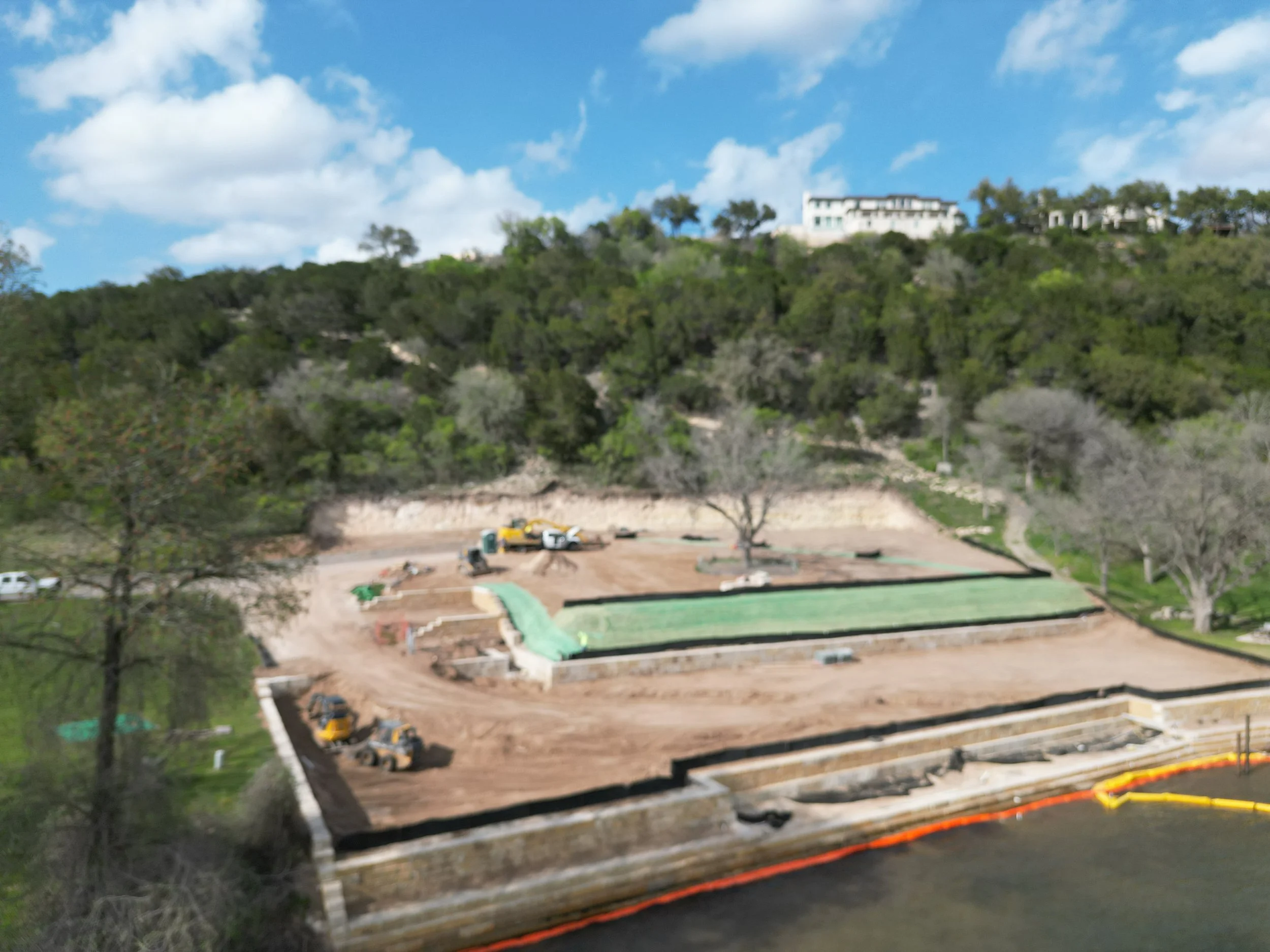 Construction site near a body of water with excavators, dirt, and green construction materials, surrounded by trees and hills.