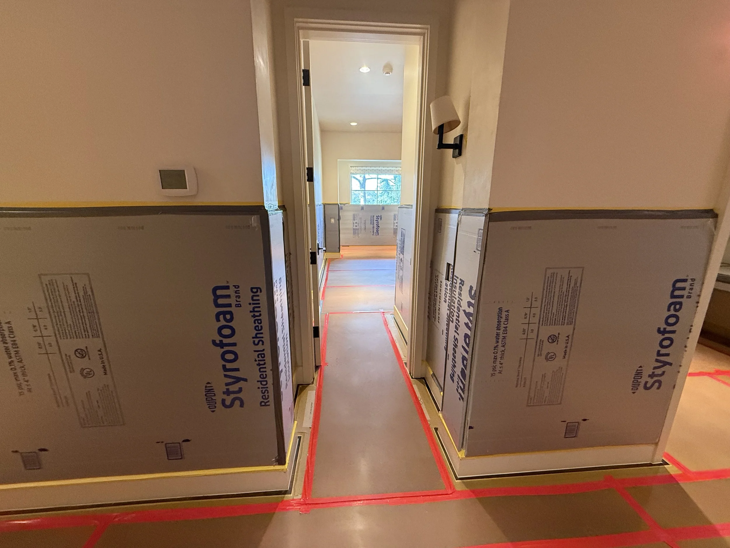Hallway under construction with large foam boards and red tape on the floor, leading to a room with a window in the background.