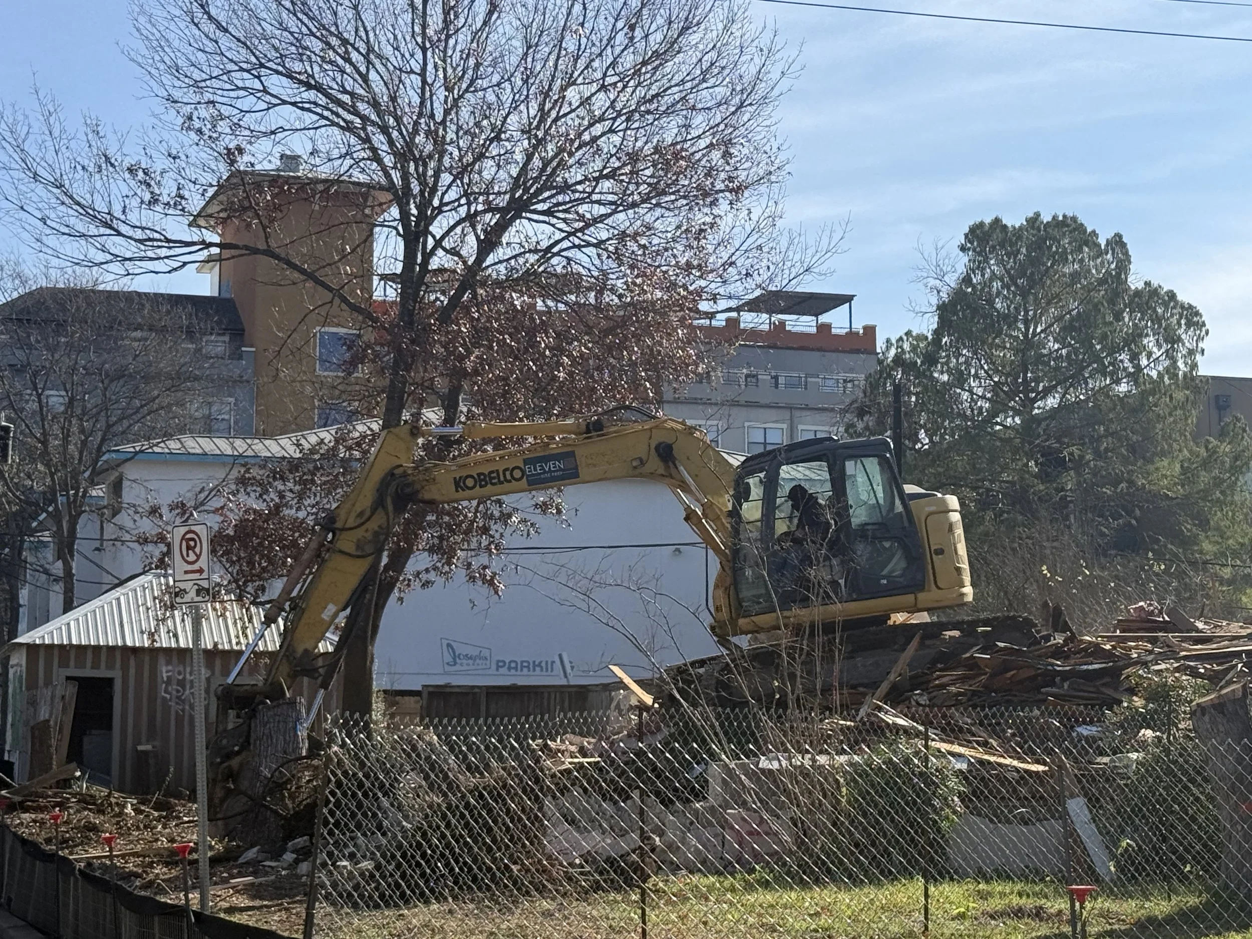 Excavator demolishing a small wooden structure in an urban area with trees and multi-story buildings in the background.
