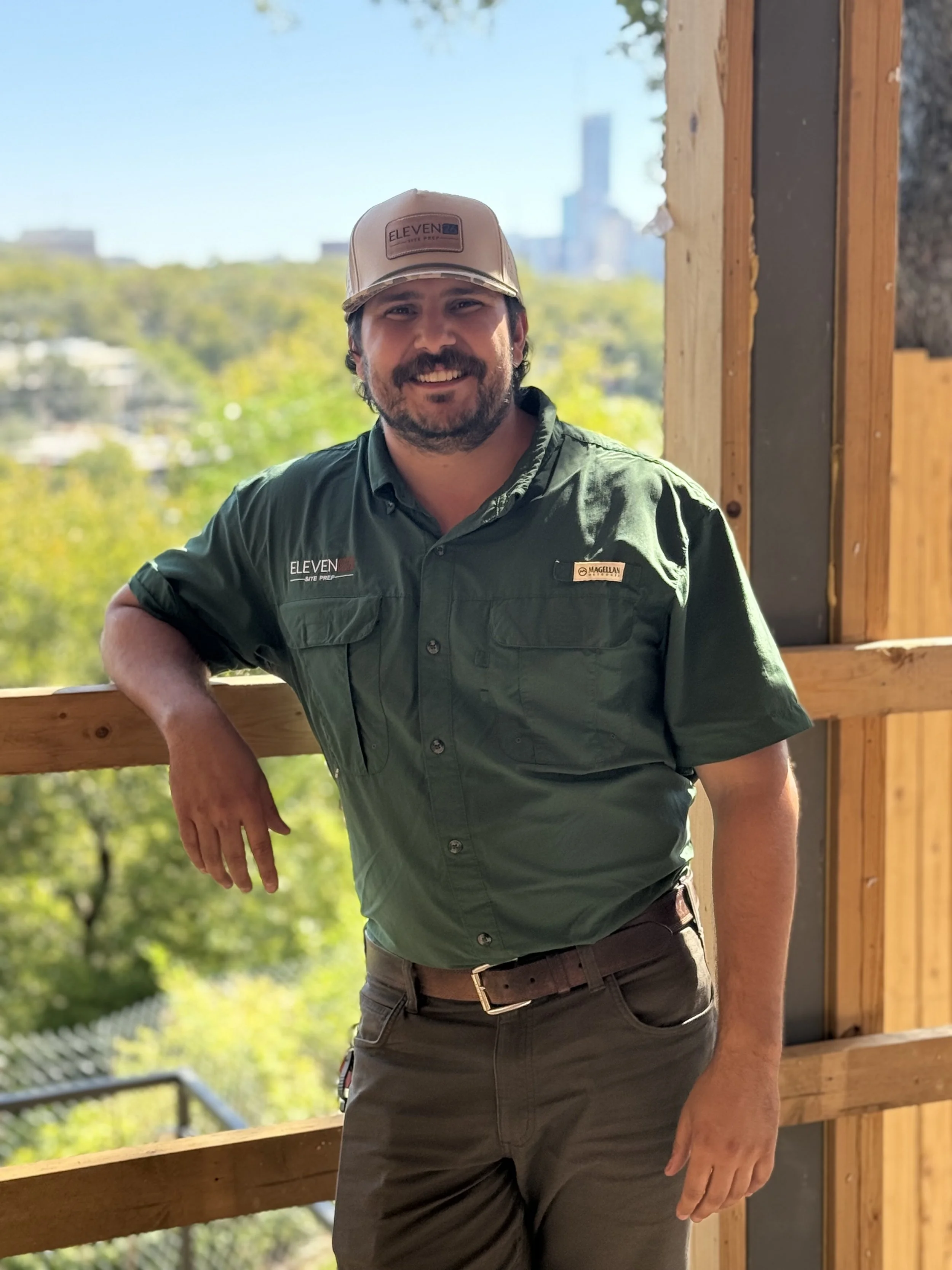 Man with beard wearing a green shirt and a beige cap, standing on a construction site with wooden framing, overlooking trees and city buildings in the background.