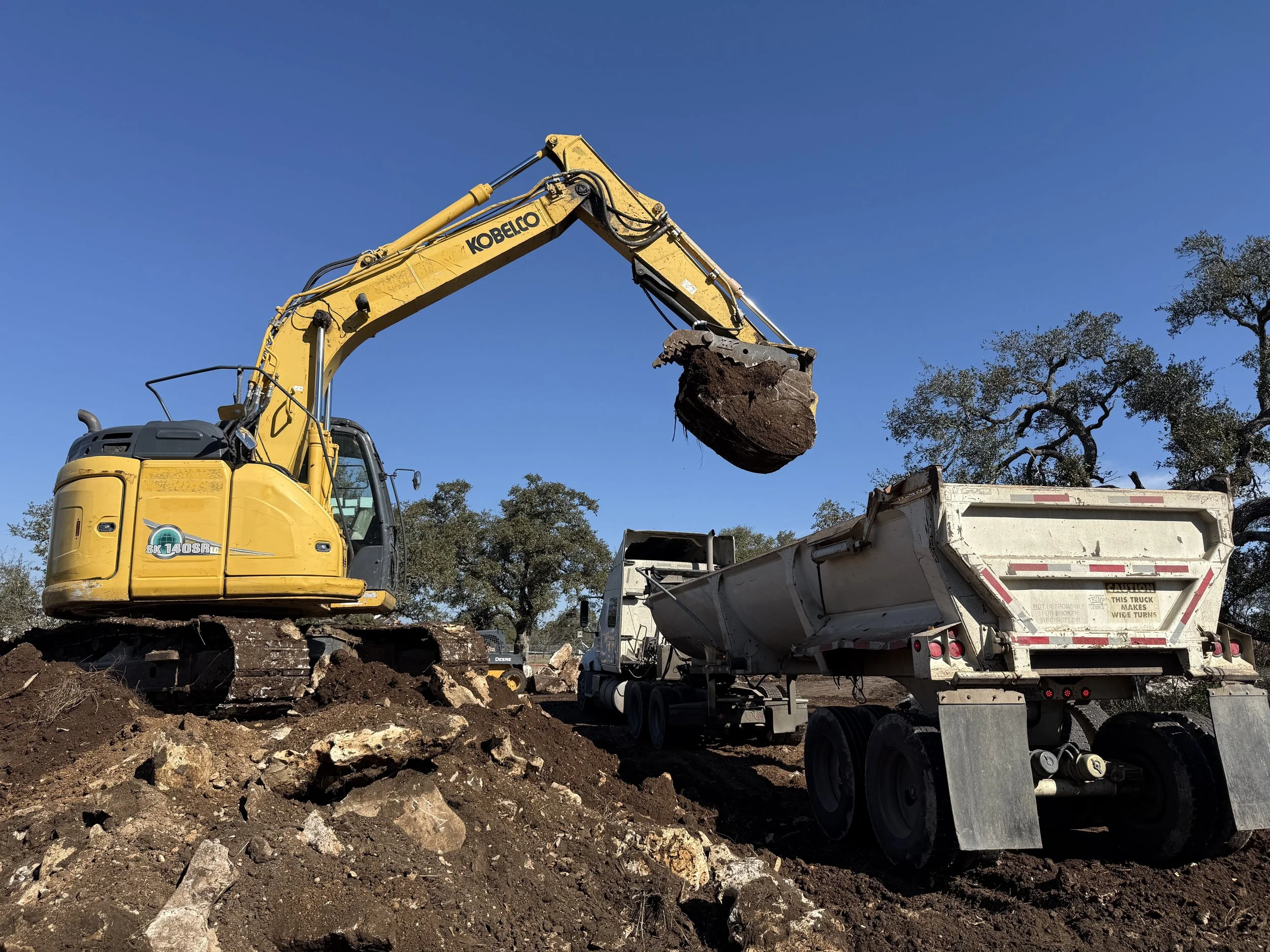 A yellow excavator loading dirt and rocks into a dump truck on a construction site under a clear blue sky with trees in the background.