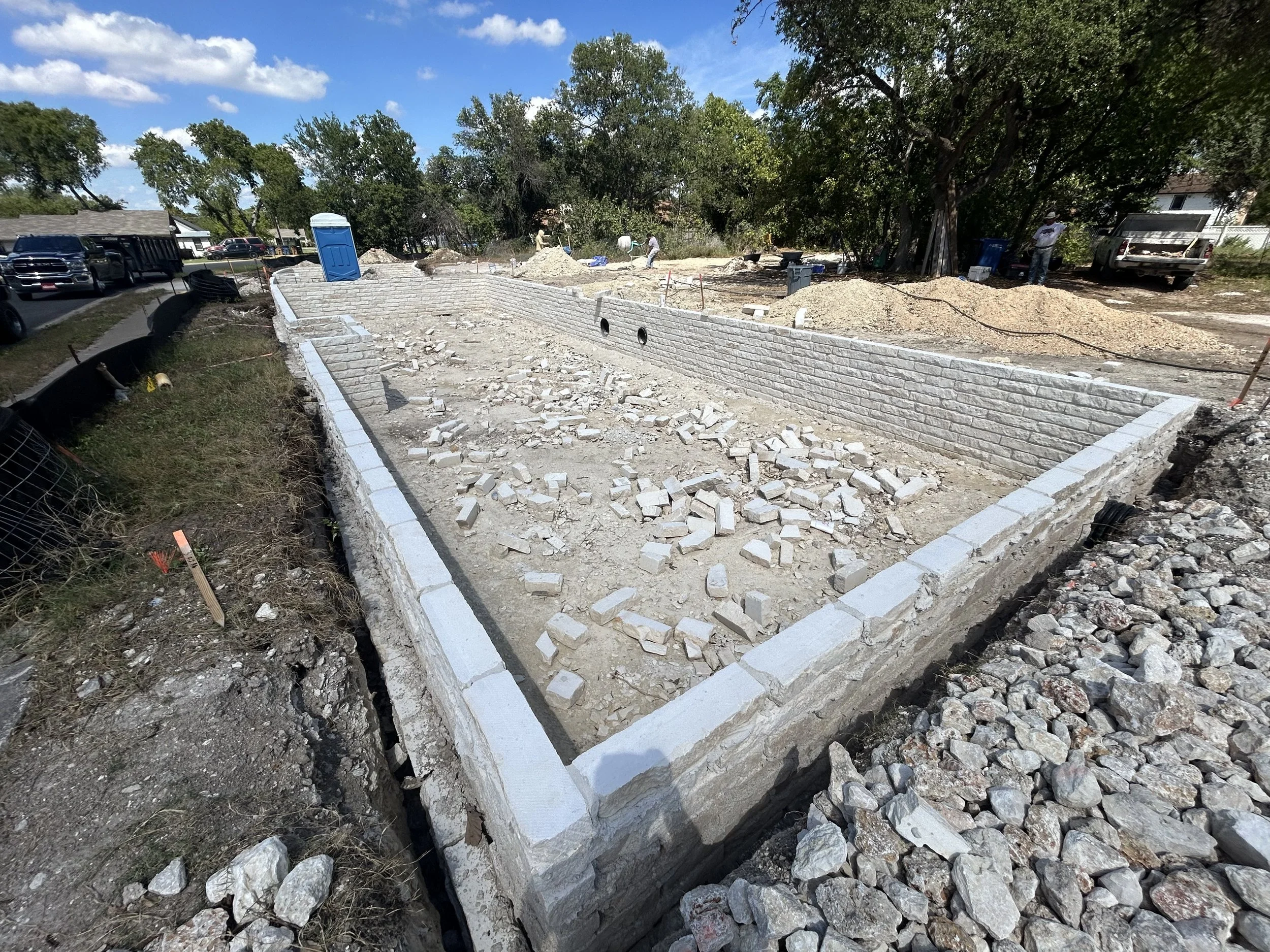 Construction site with a partially built brick wall, rubble on the ground, surrounded by trees and parked cars.