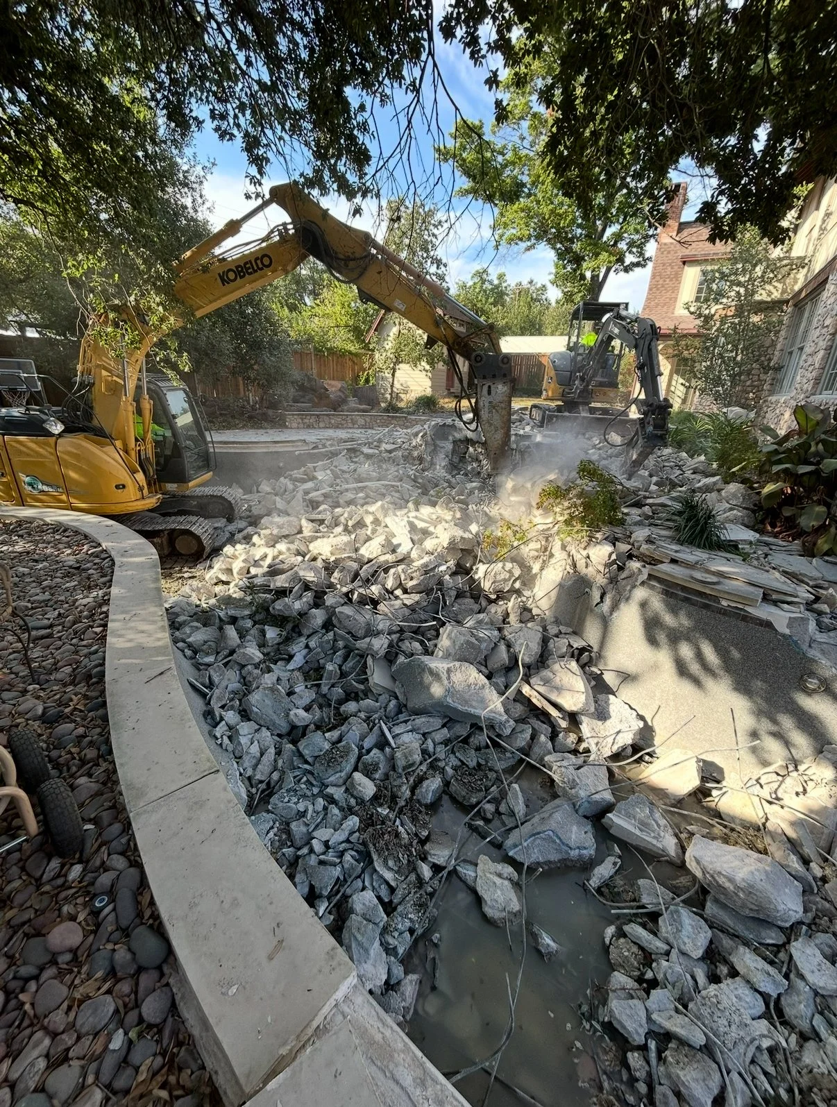 Construction site with two excavators demolishing a concrete swimming pool filled with rubble and debris, with trees and houses in the background.