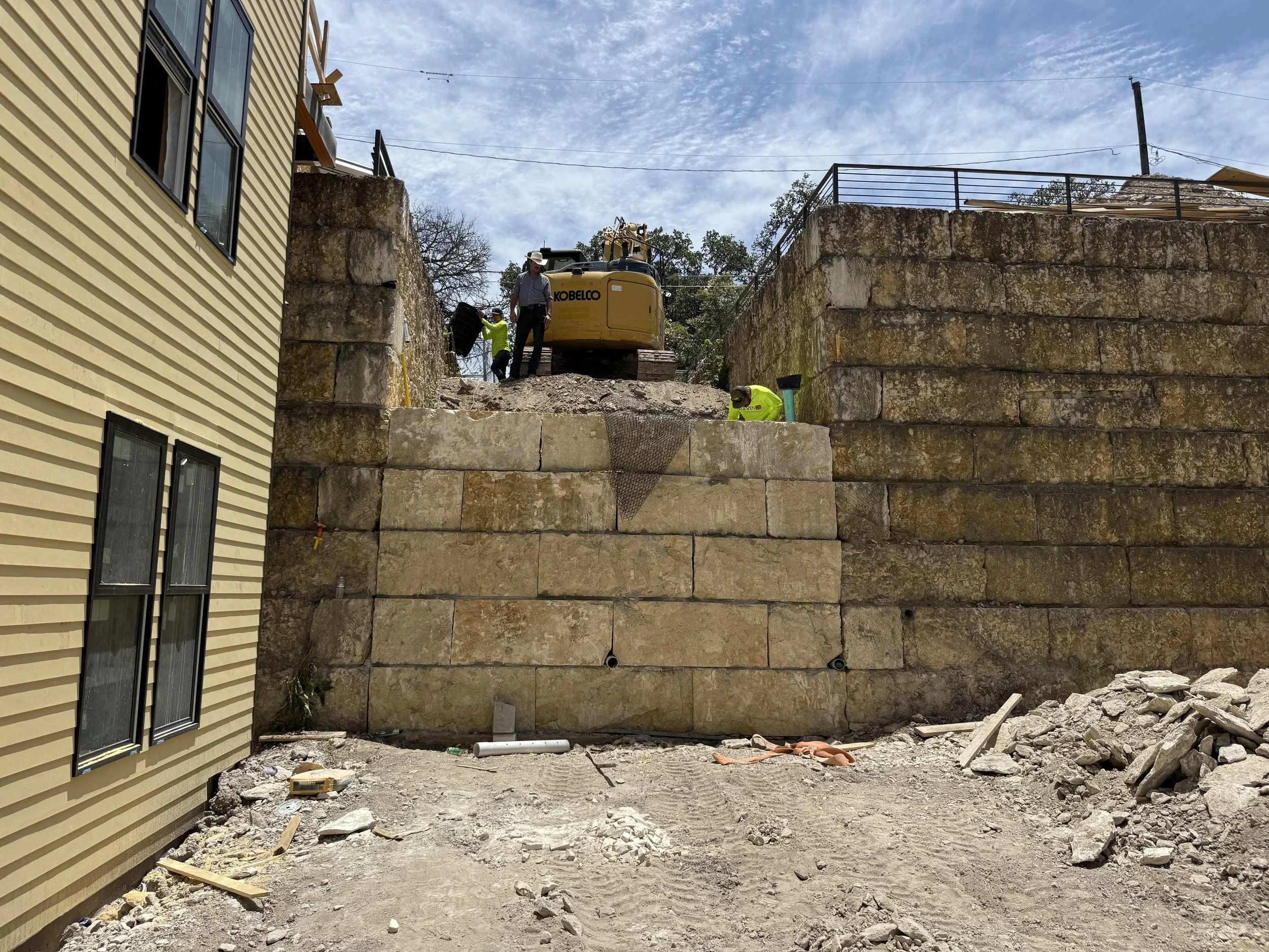 Construction workers building a retaining wall with large stones on a sloped terrain, with a yellow Kobelco excavator nearby. The site is between a yellow house and a stone-paved area, under a partly cloudy sky.