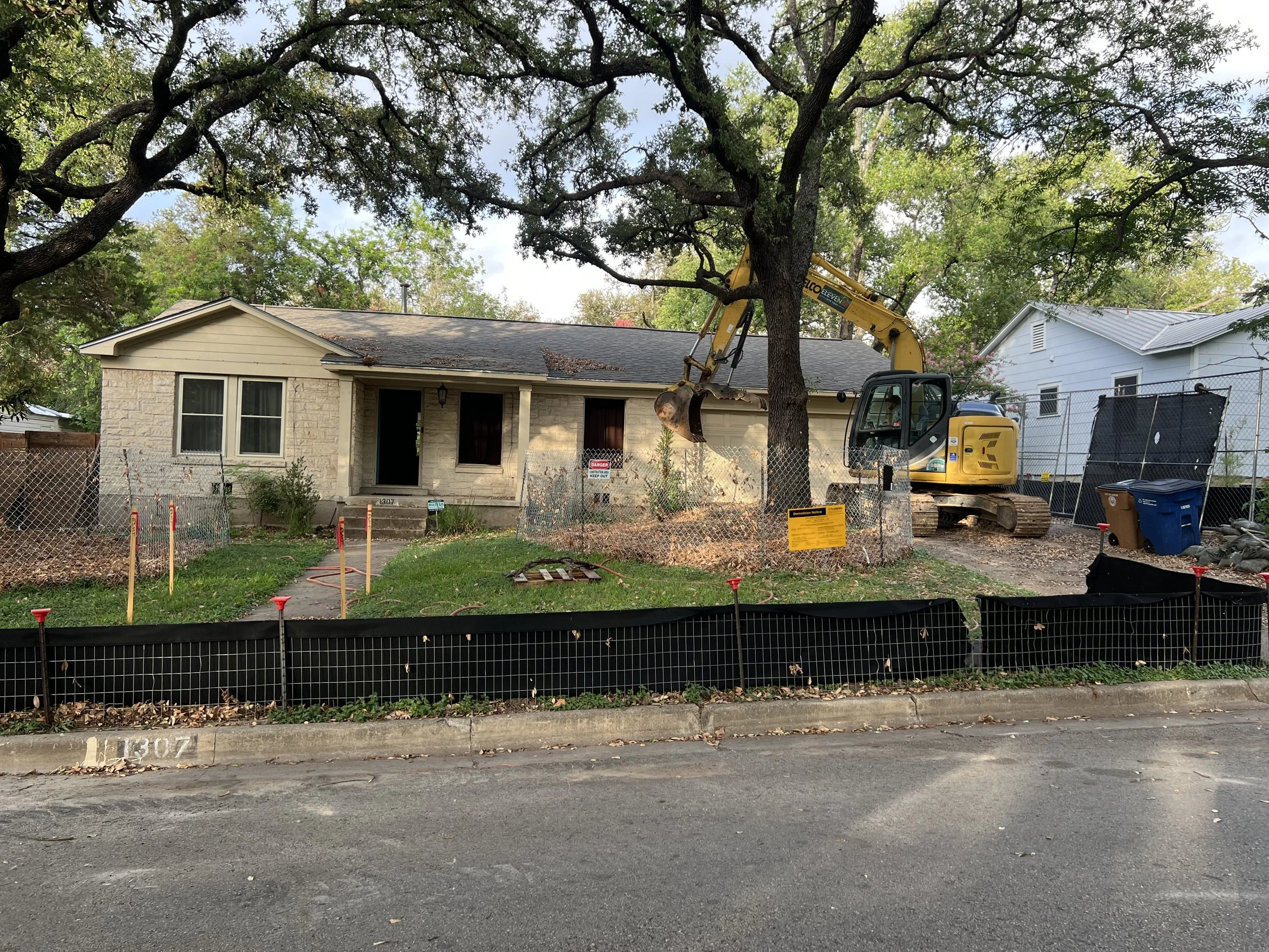 A house with a tree in front, surrounded by construction fencing and a small excavator removing part of the tree.