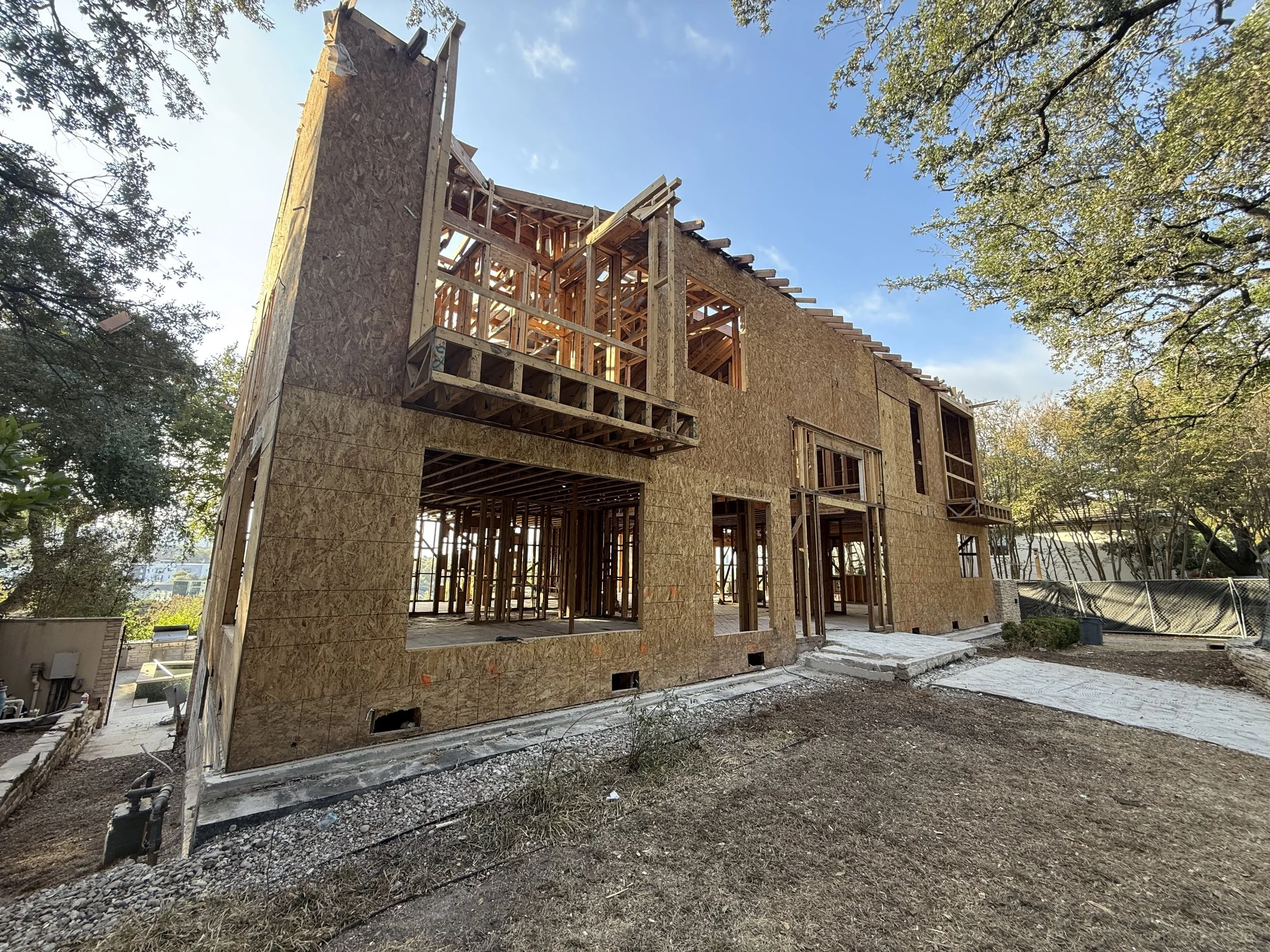 Under-construction two-story house with visible wooden framing and sheathing panels, surrounded by trees on a clear day.