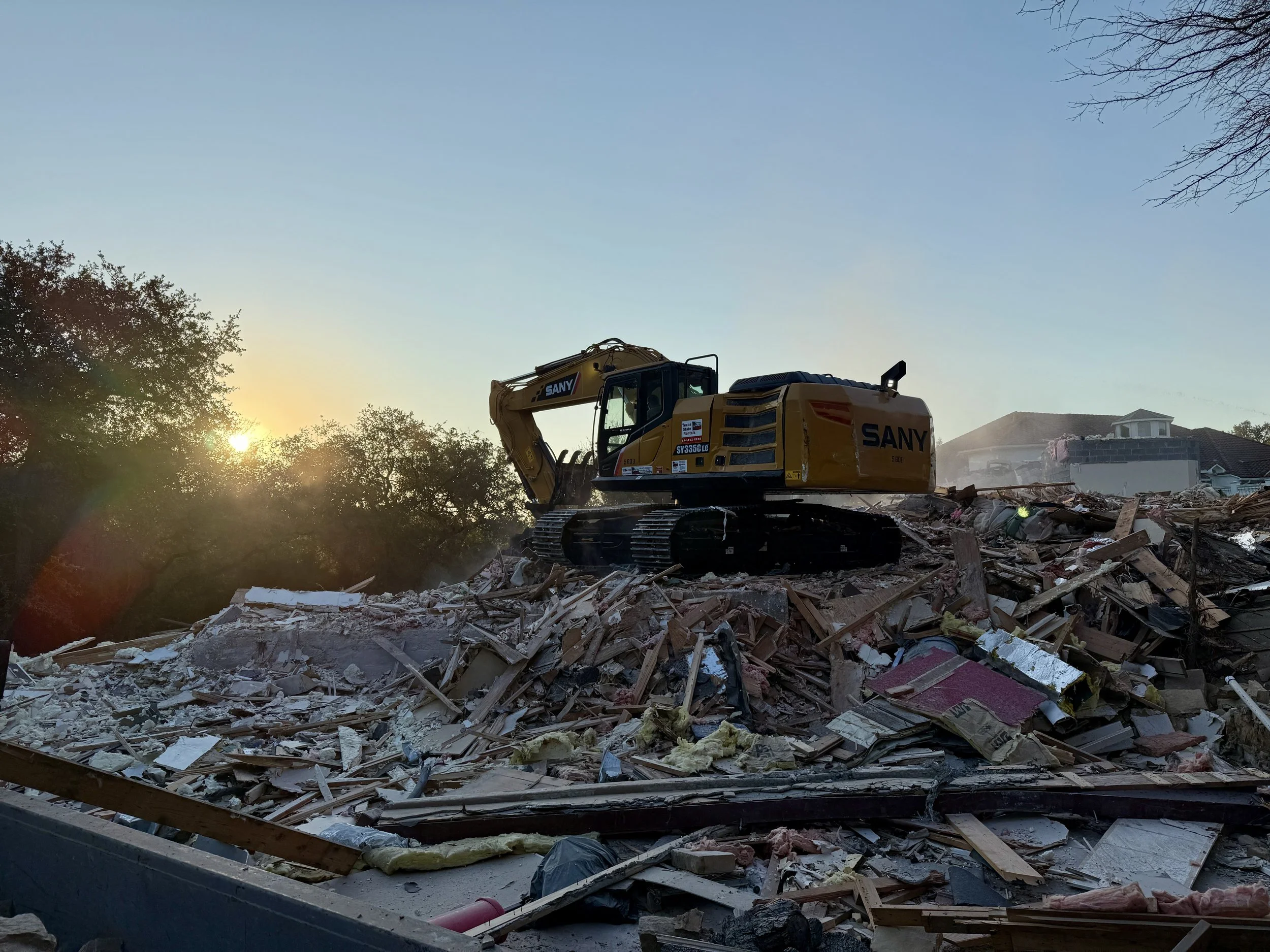 A yellow SANY excavator working amid rubble and debris of demolished buildings at sunrise.