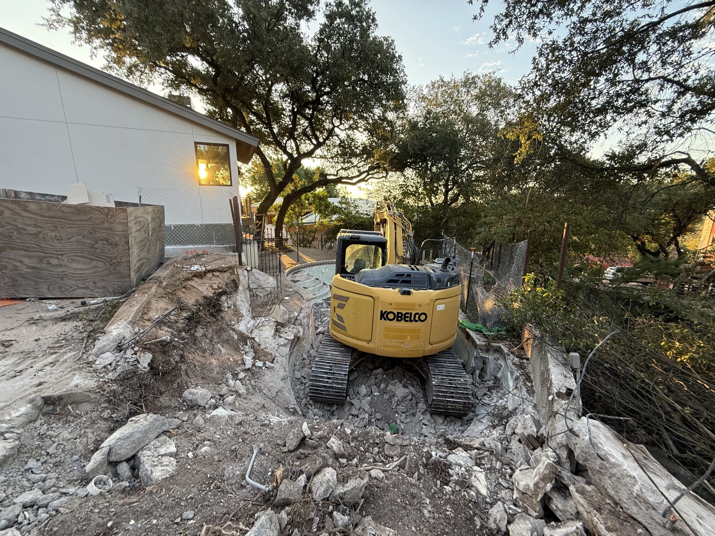 Construction site with a Kobelco mini excavator on uneven dirt ground, surrounded by rocks, dirt piles, and construction fencing, with trees and a white building with a window reflecting sunlight in the background.