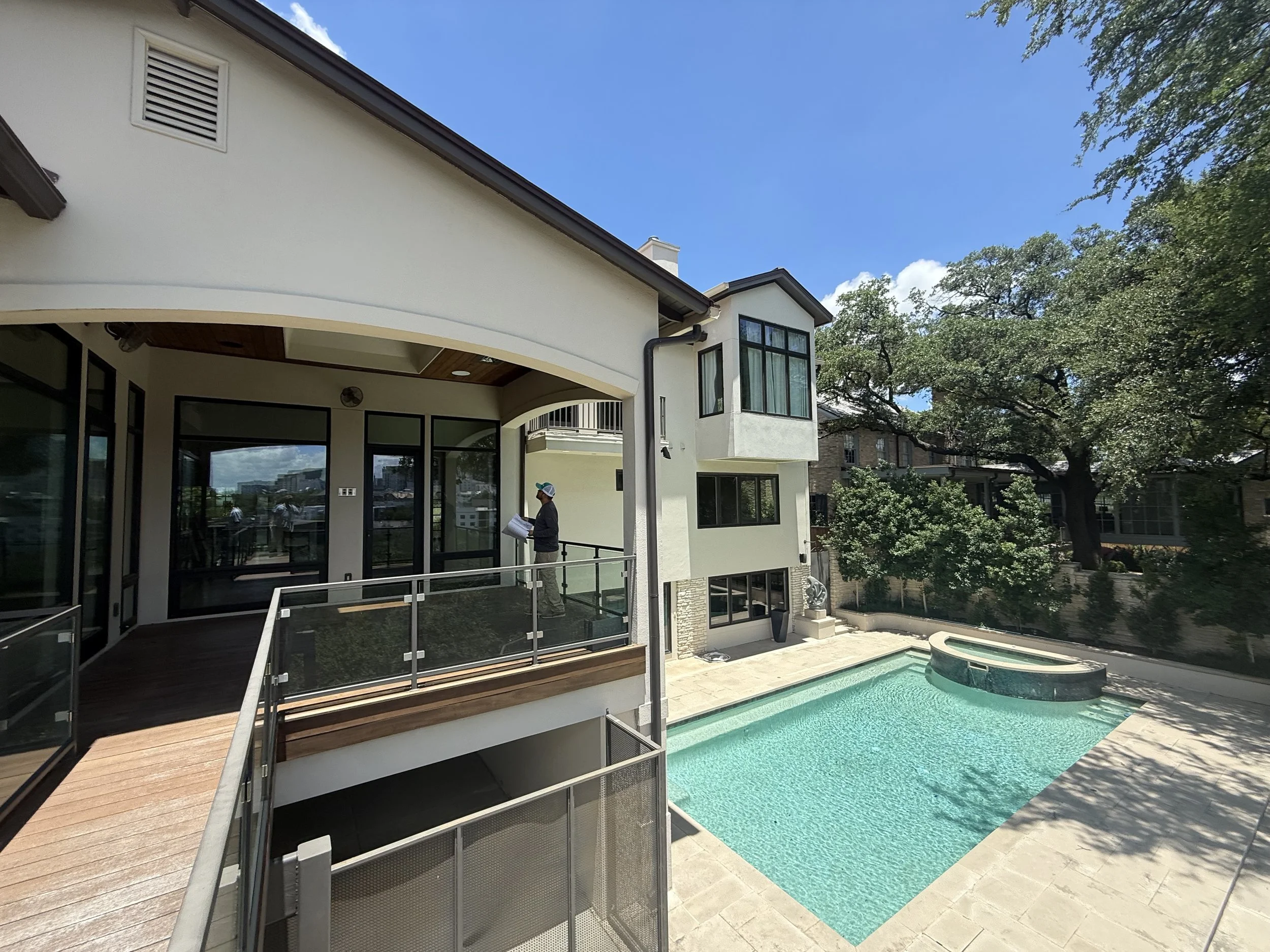 A modern two-story house with large glass windows, a swimming pool, and an outdoor deck with a person inspecting blueprints.