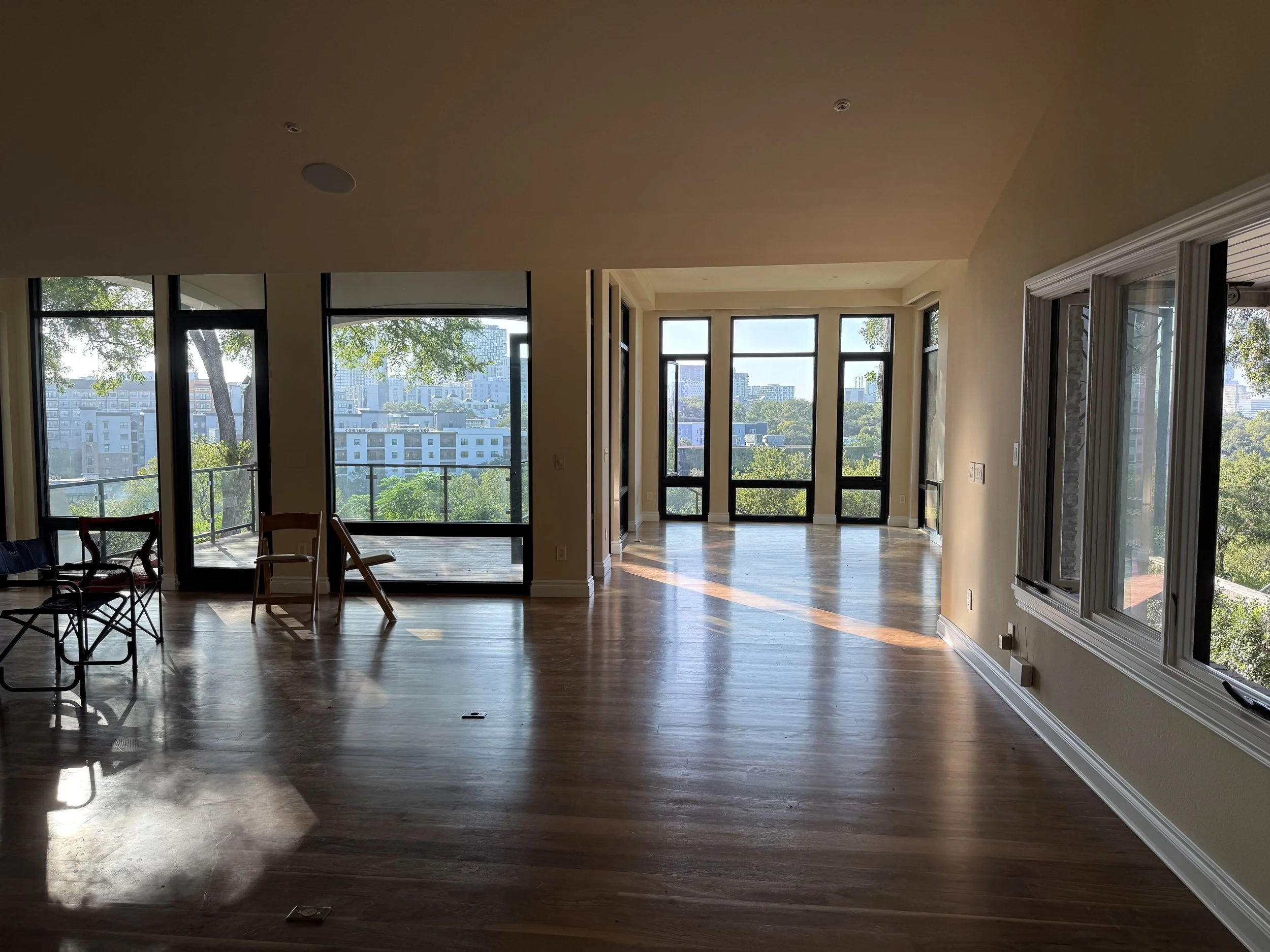 Empty living room with large windows and hardwood floors, sunlight streaming in, and some chairs near a balcony with a city view.