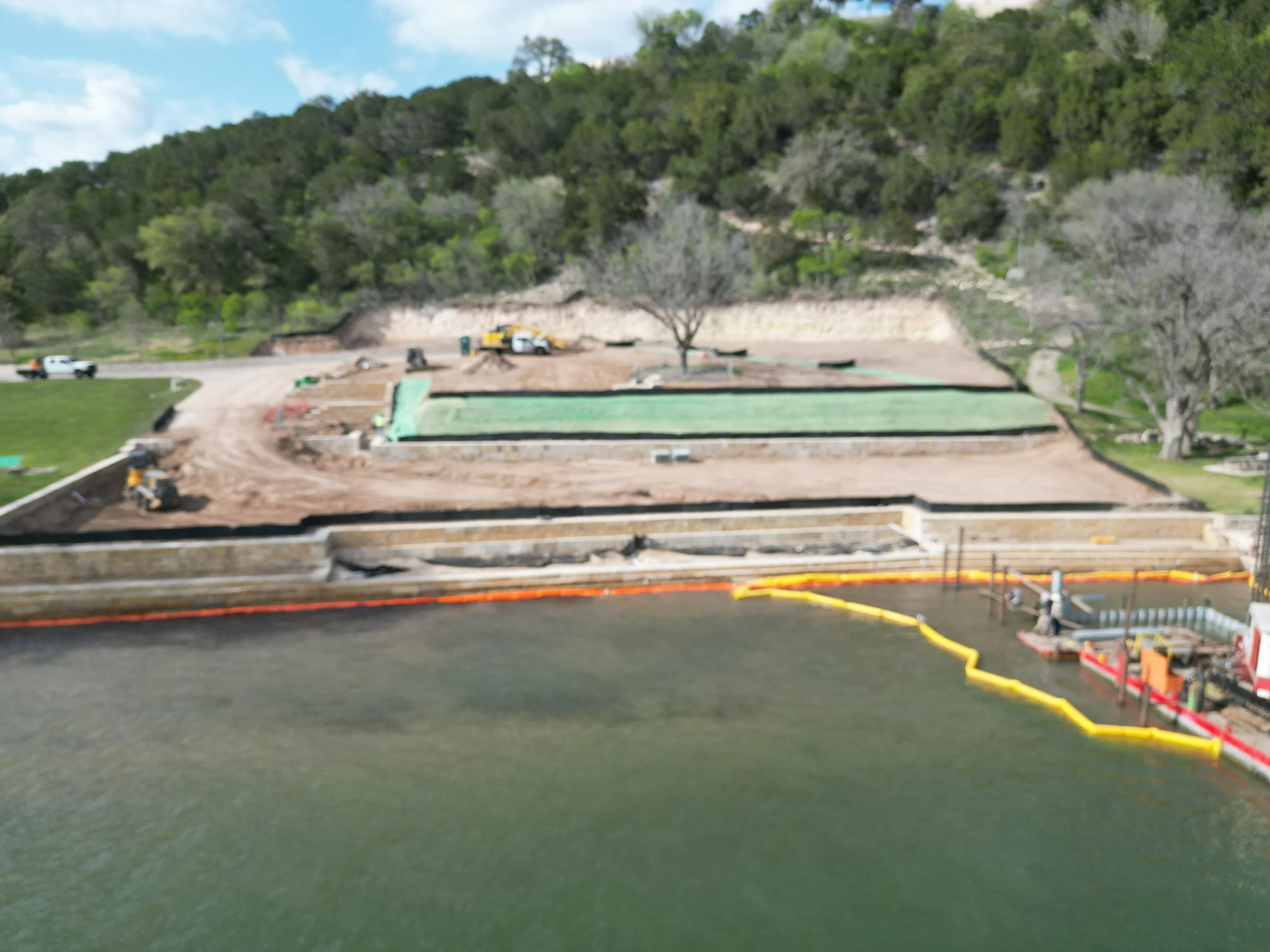 Construction site by a body of water with multiple landscaped tiers, construction vehicles, and trees in the background.