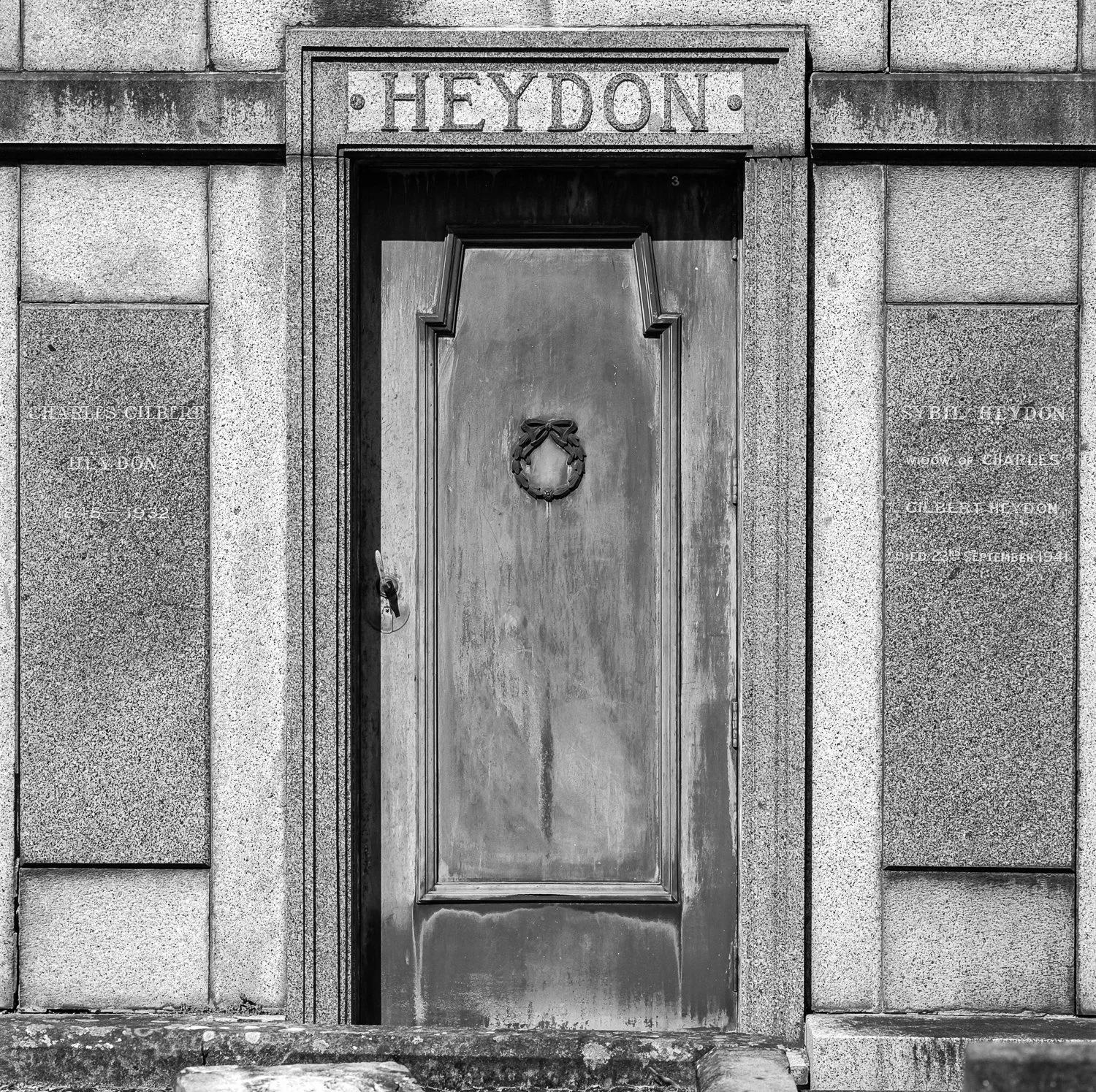 Black and white photo of a historic stone building entrance with a wooden door, wreath decoration, and inscriptions on plaques on either side of the door, including the name 'Heydon' at the top.