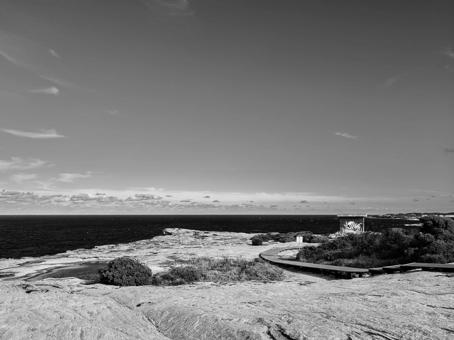 Black and white photograph of a coastal landscape with a rocky shoreline, sparse vegetation, a winding walkway, and a building near the water against a sky with scattered clouds.