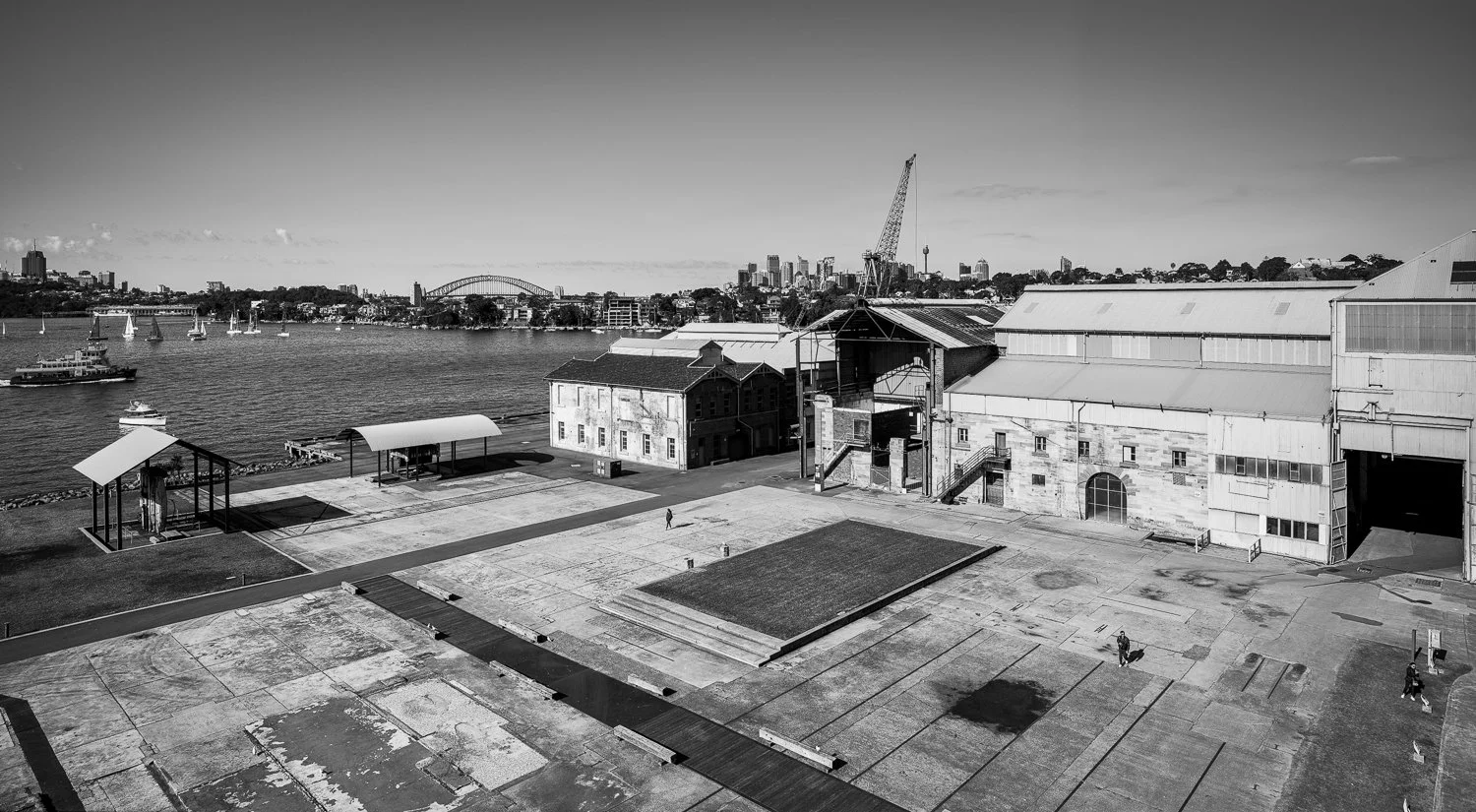A black and white photo of a waterfront scene with several boats on the water, industrial buildings, and a city skyline with tall buildings in the background.