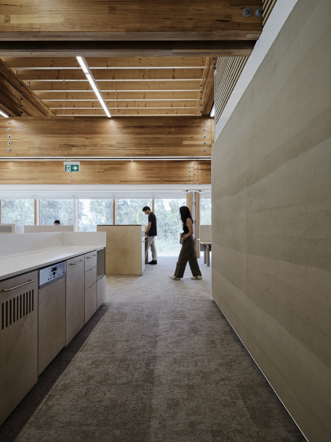 Interior of a modern room with wooden ceiling and light-colored walls, featuring a hallway with people walking and large windows in the background.