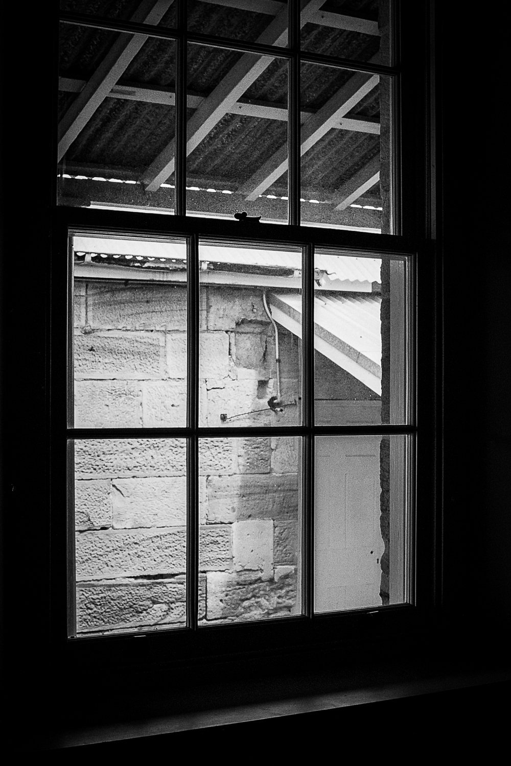Black and white photo of a multi-pane window with a view of an outdoor wall made of bricks and exposed beams of a roof.