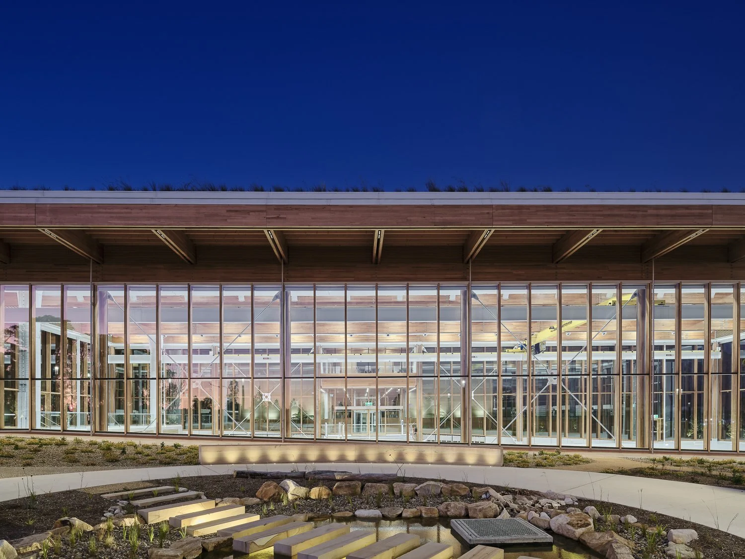 Modern glass-front building with wooden roof structure, illuminated interior, and landscaped exterior courtyard with rocks, plants, and stepping stones at night.
