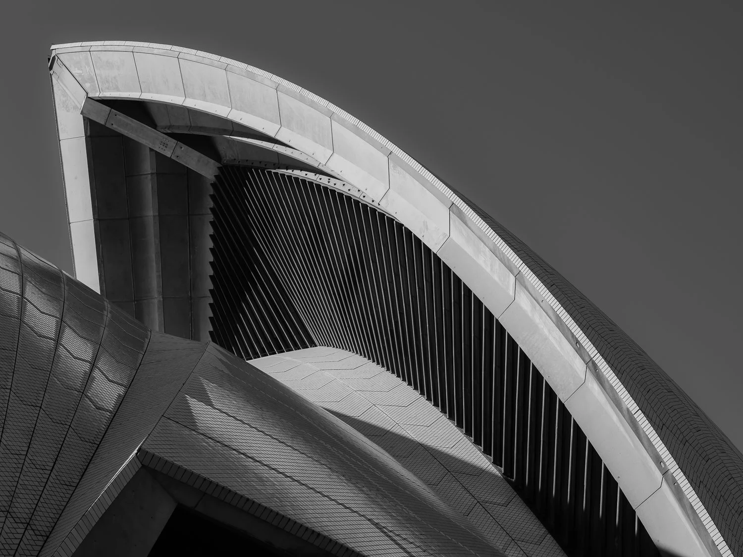 Close-up of a modern architectural building with curved metallic surfaces and vertical slats, viewed from below against a gray sky.