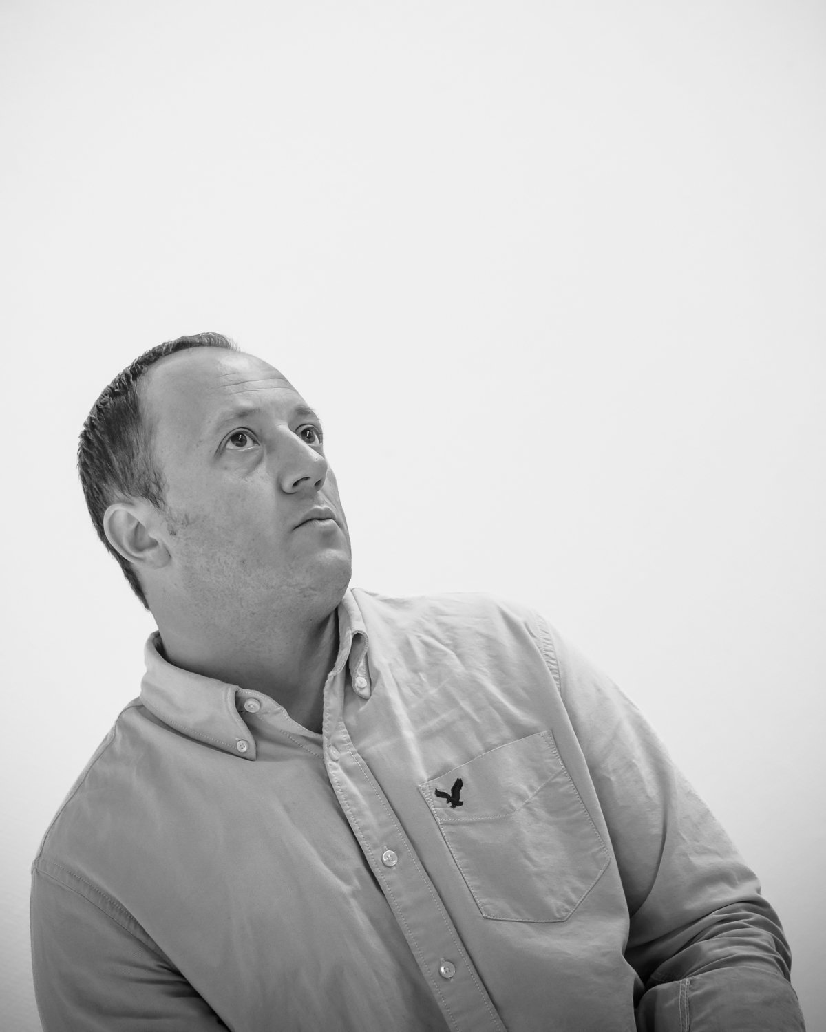 Black and white photo of a man looking upward with a neutral expression, wearing a button-up shirt with a small bird emblem on the pocket, standing against a plain background.