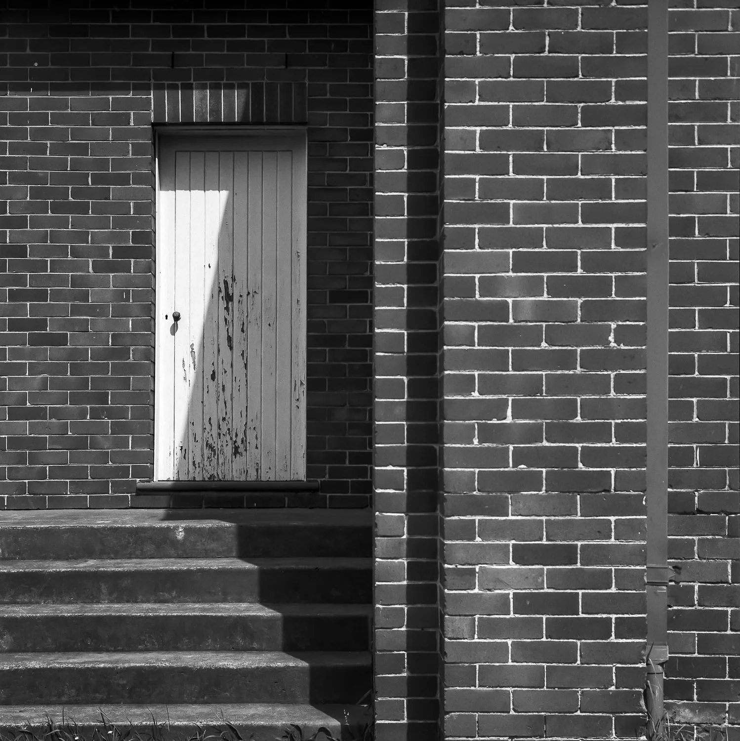 Black and white photo of a brick building with a weathered wooden door and steps leading up to it, casting a shadow.