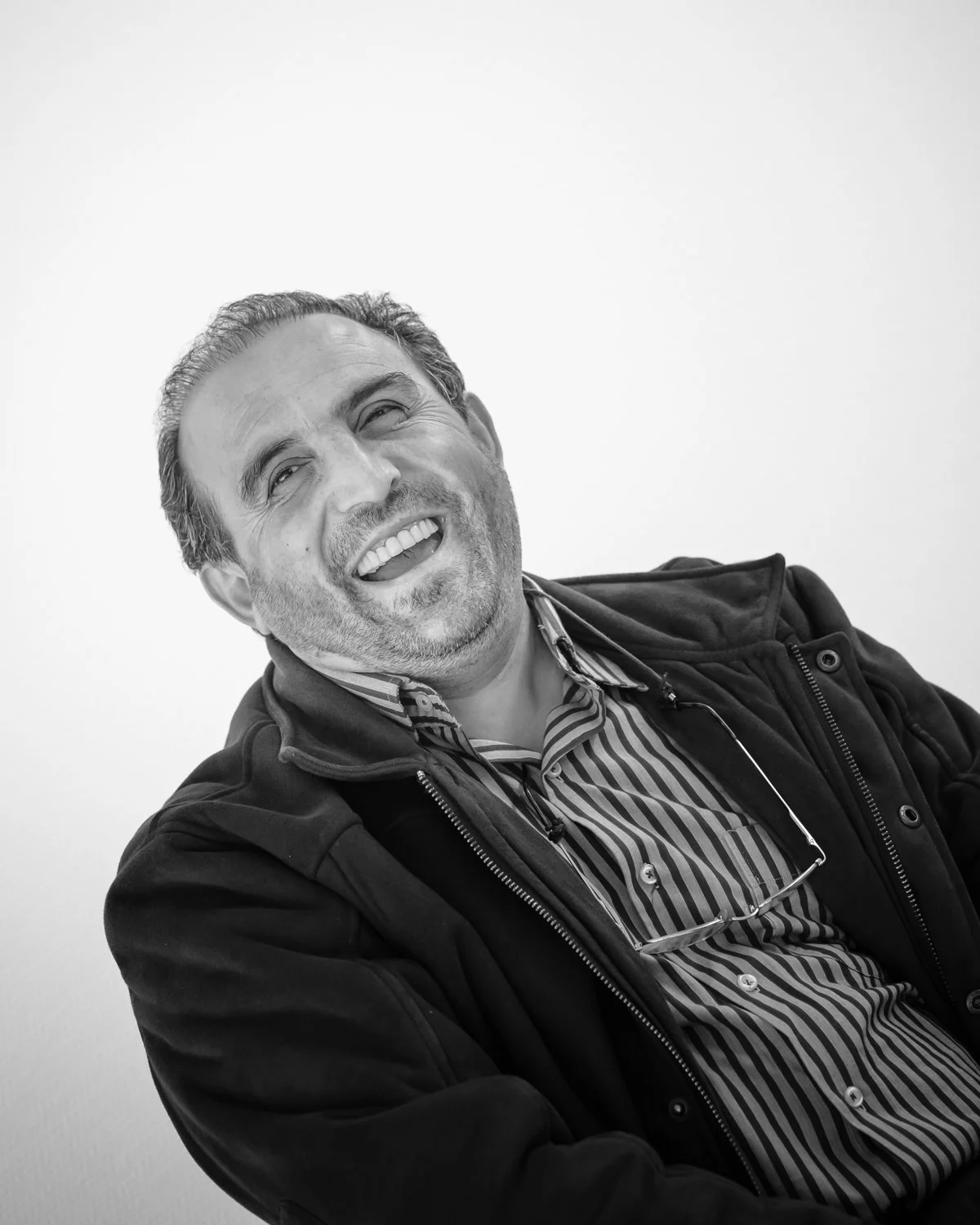 A smiling man with short curly hair, wearing a striped shirt and a dark jacket, sitting against a plain background.
