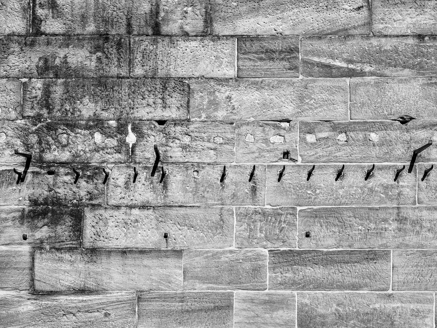 A black and white photo of a brick wall with multiple hooks attached to it, some of which are bent or missing.
