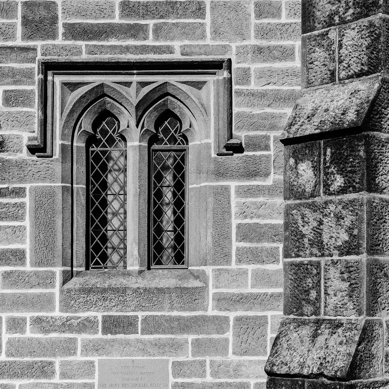 Black and white photo of a stone church wall with a stained glass window featuring a pointed gothic arch and diamond-shaped leaded glass panes.
