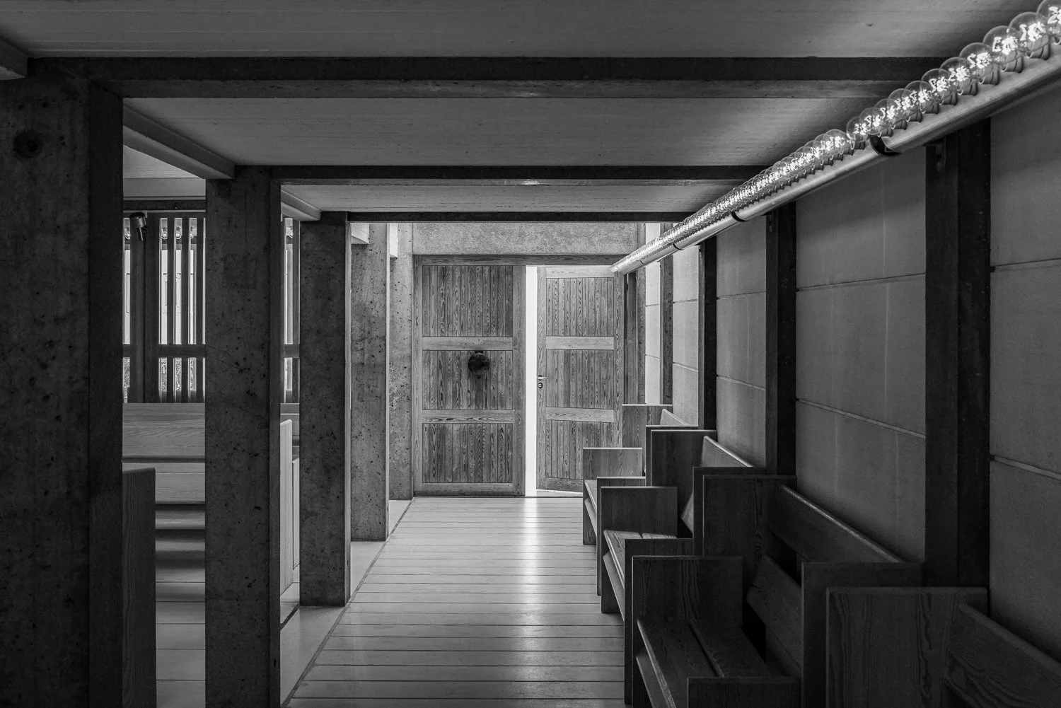 Black and white photo of a wooden hallway in a modern building with benches along the wall and a box-type ceiling.