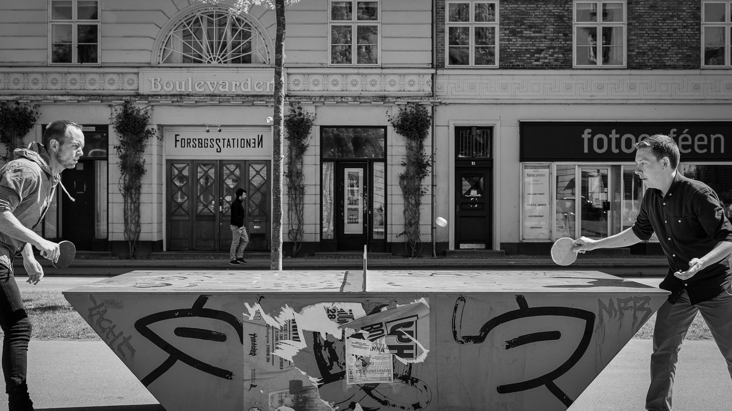 Two people playing ping pong on an outdoor table in front of a street with buildings and pedestrians in the background.