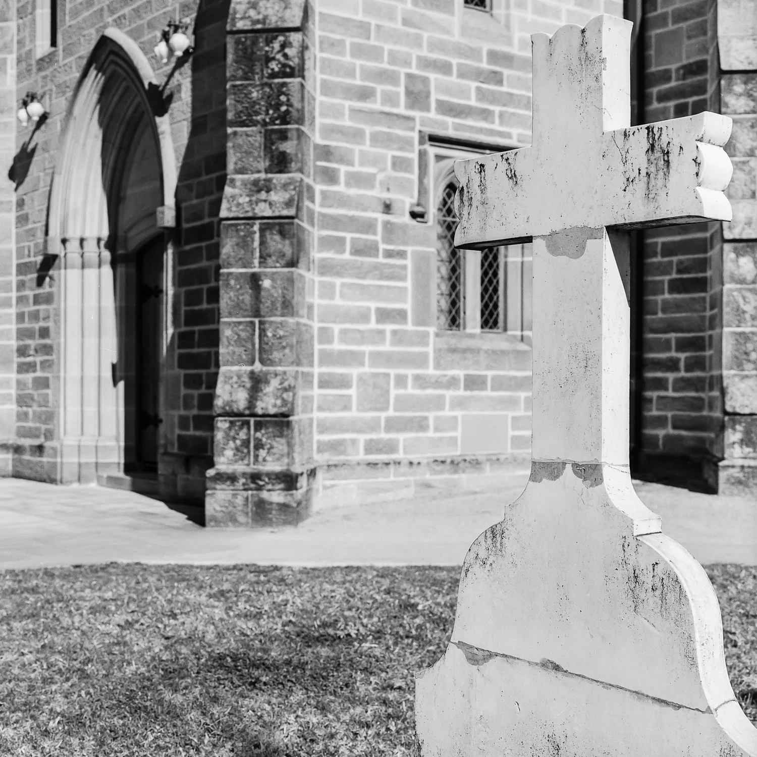 A stone cross monument in front of a brick church building with arched doorway and small windows.
