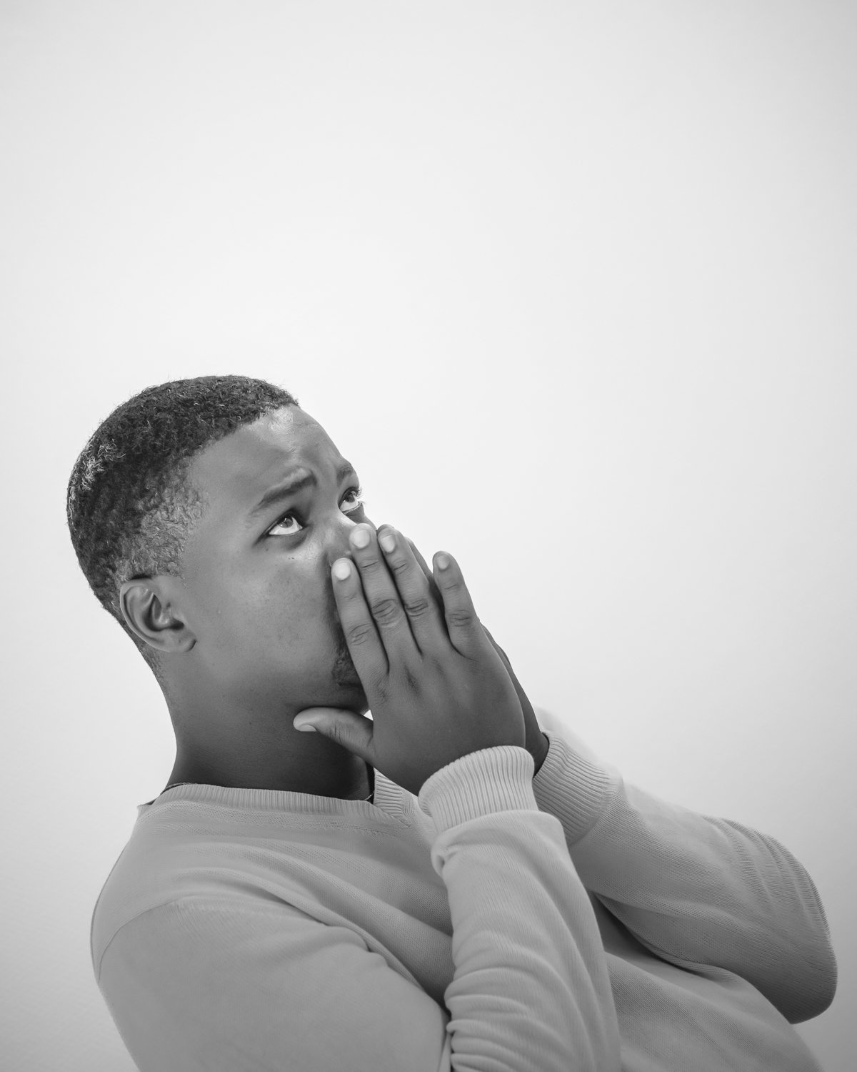 A young man with short hair looking up with a worried or anxious expression, covering his mouth with both hands.