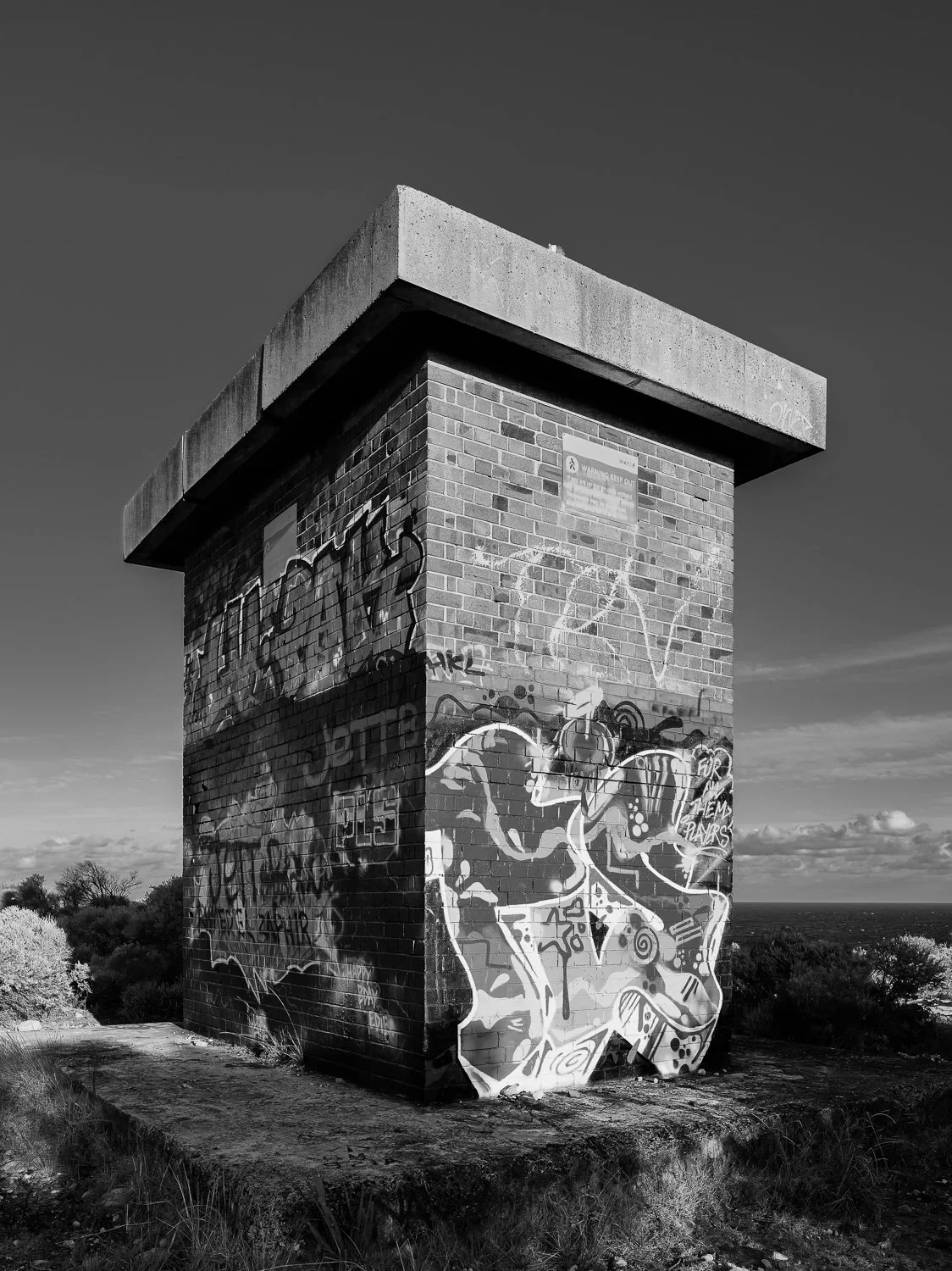A brick tower with graffiti and art on its walls, set in an outdoor landscape with trees and a cloudy sky in the background.