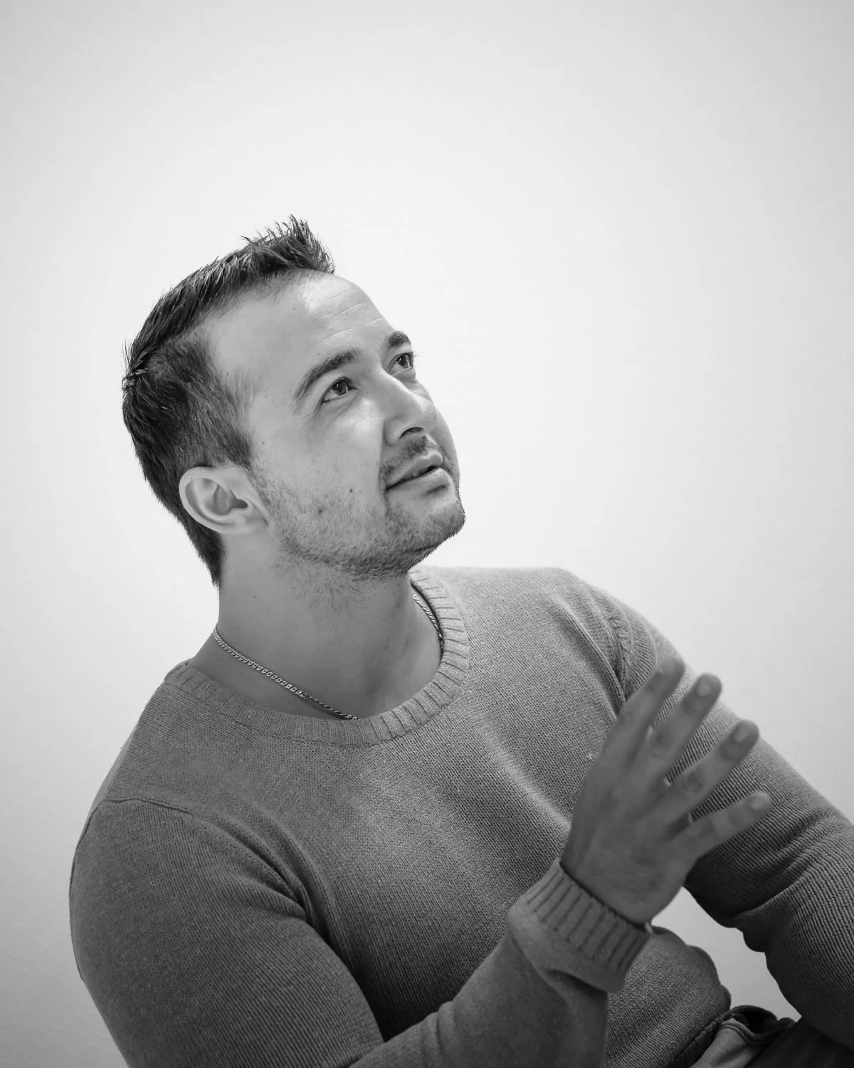 Black and white photo of a young man with short dark hair and a beard, gesturing with his hand while looking upward, wearing a sweater.