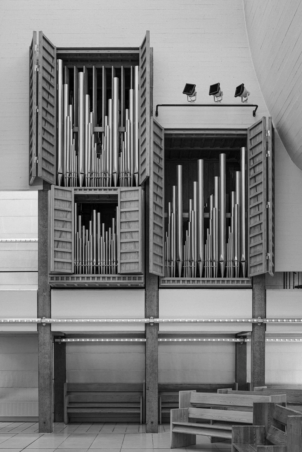 Large pipe organ with multiple pipes in wooden cabinets, located inside a building with white walls and overhead lighting.