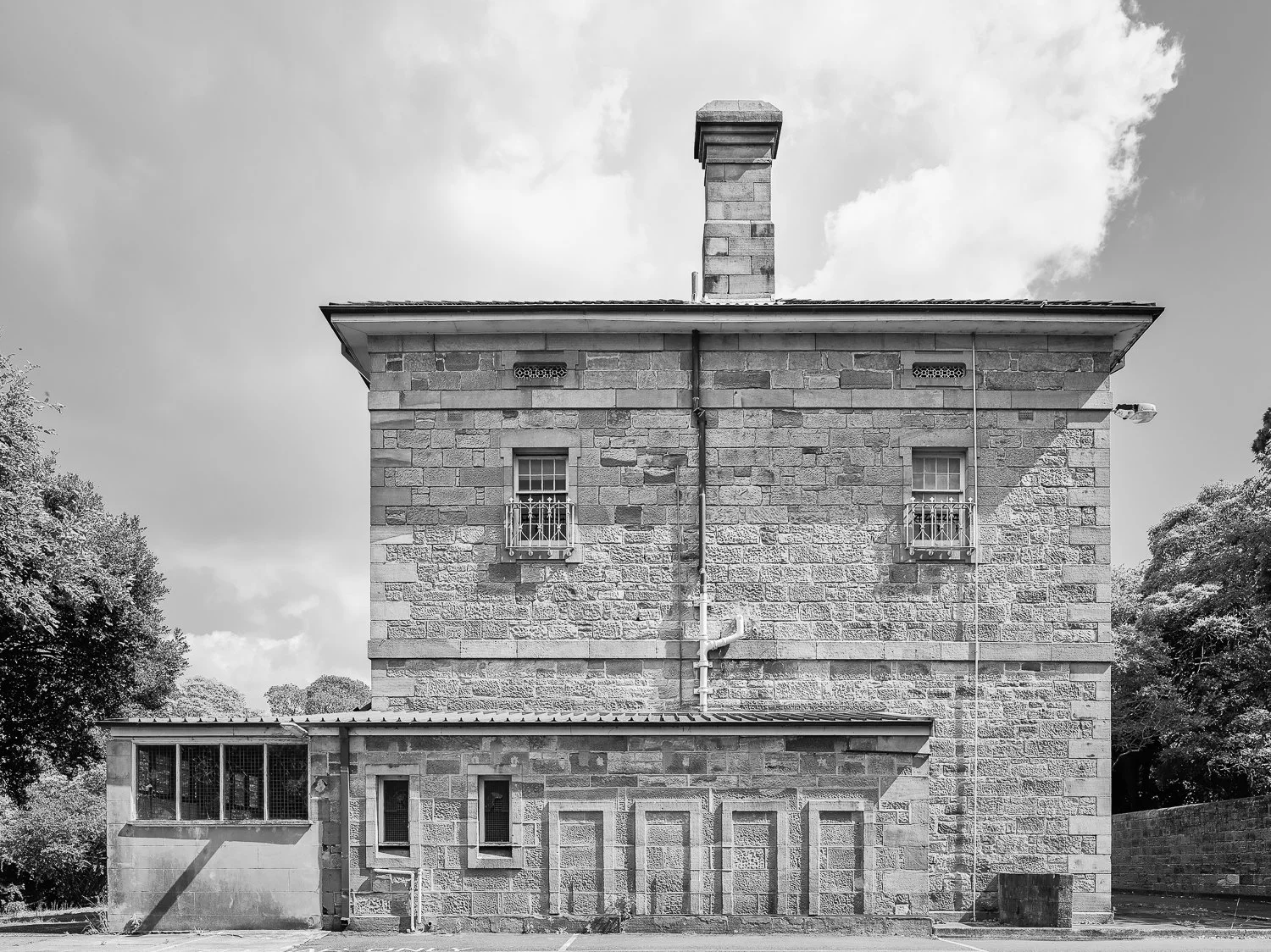 Black and white photo of a two-story stone house with a chimney, barred windows, and a small side building with three windows. Trees are visible on both sides.