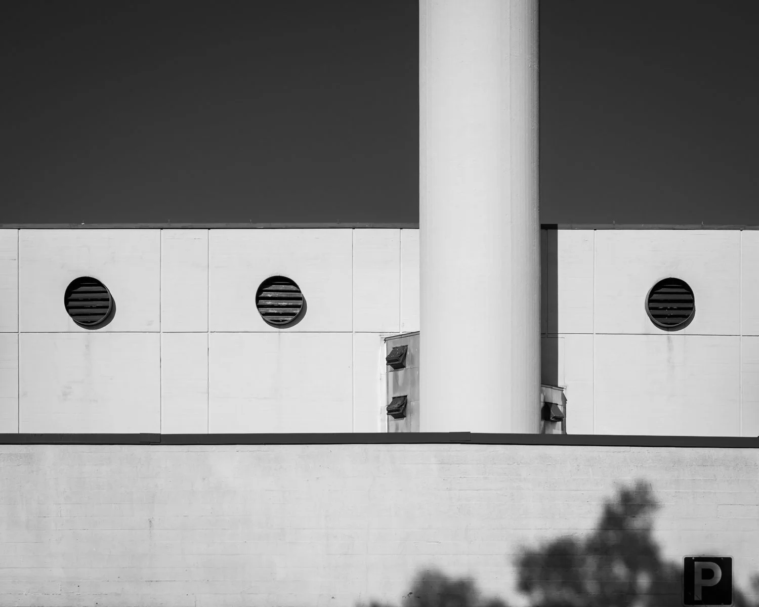 Black and white photo of a modern building with three circular vents and a large vertical pipe, with a shadow of a tree at the bottom right corner.