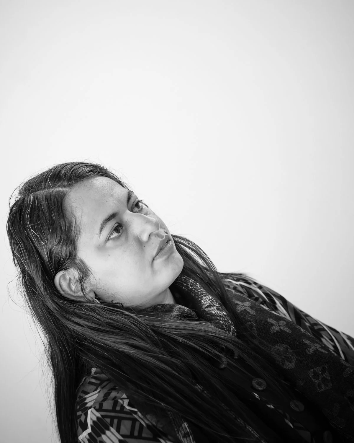 A black and white portrait of a young woman with long dark hair, looking upwards and to the left, wearing patterned clothing against a plain background.