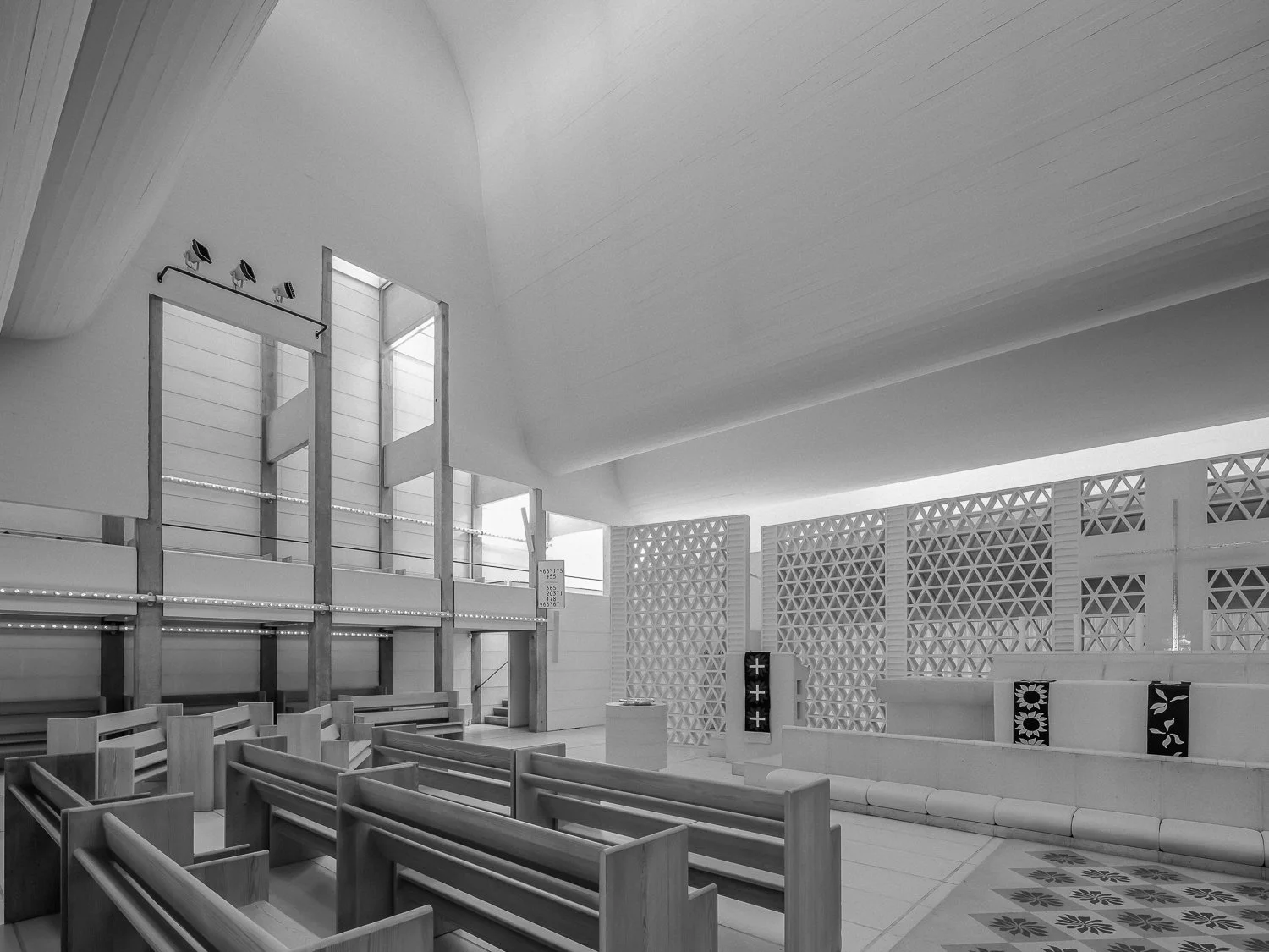 Interior of a modern church with wooden pews, decorative walls with geometric patterns, and large windows letting in natural light.