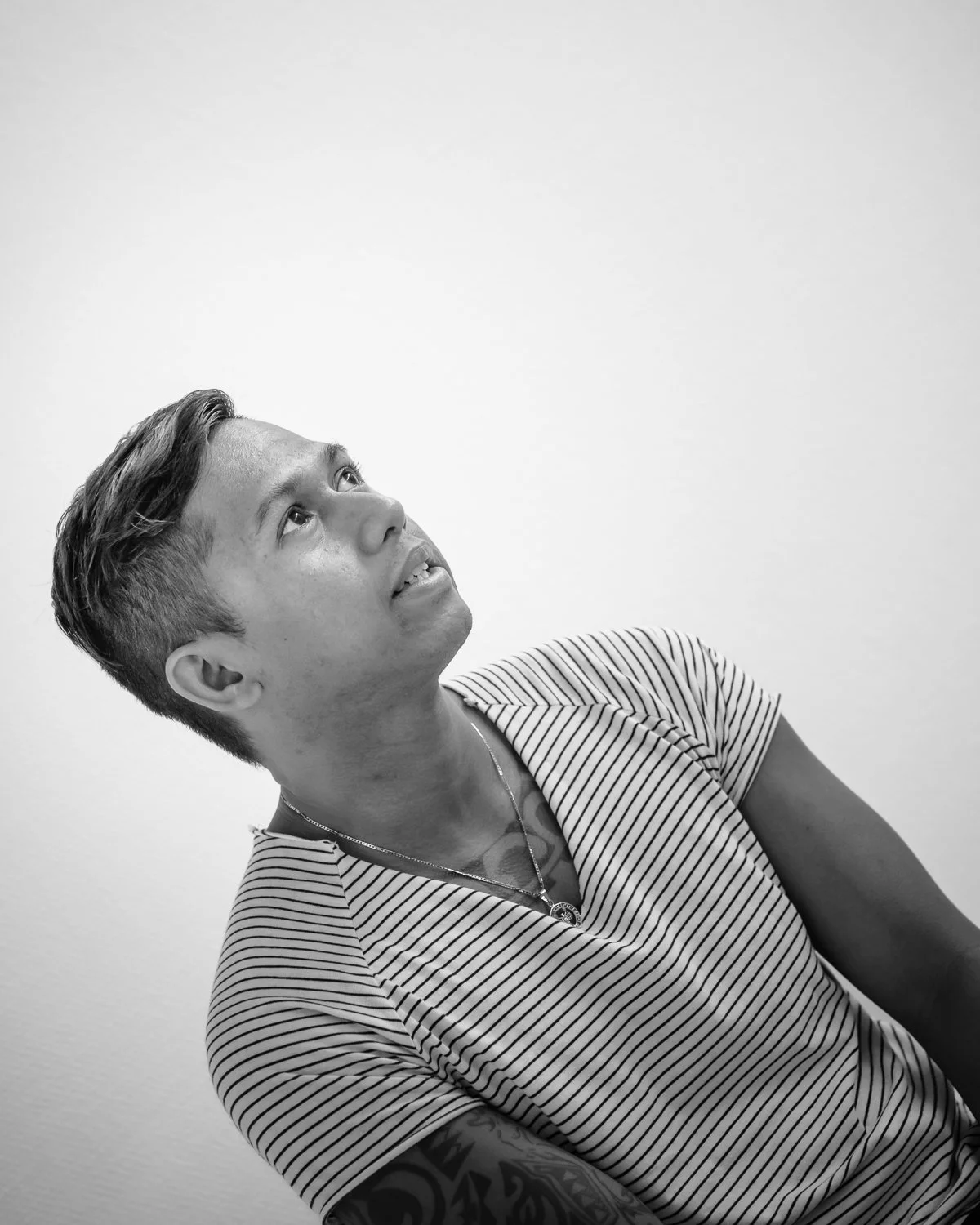 Black and white photo of a person with short hair, striped shirt, and tattoos, looking upward.