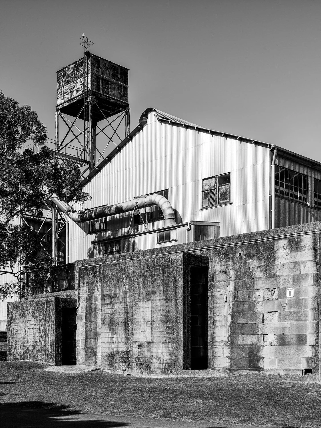 Black and white photo of an industrial building with a weathered water tower on top, a large pipe running across the side, and a concrete wall in the foreground.