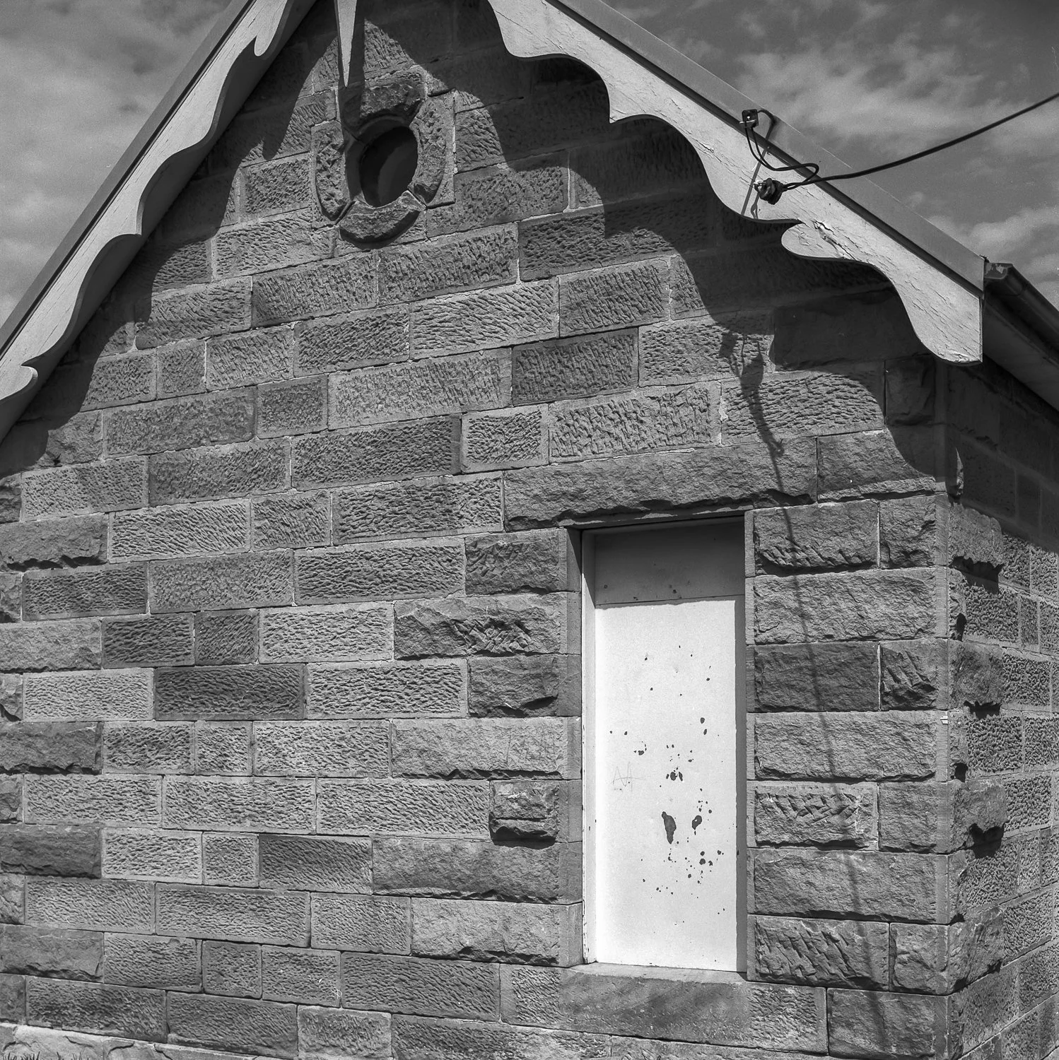 A black and white photo of a stone building with an empty window frame and a small circular opening near the gable roof, which projects above the main wall.
