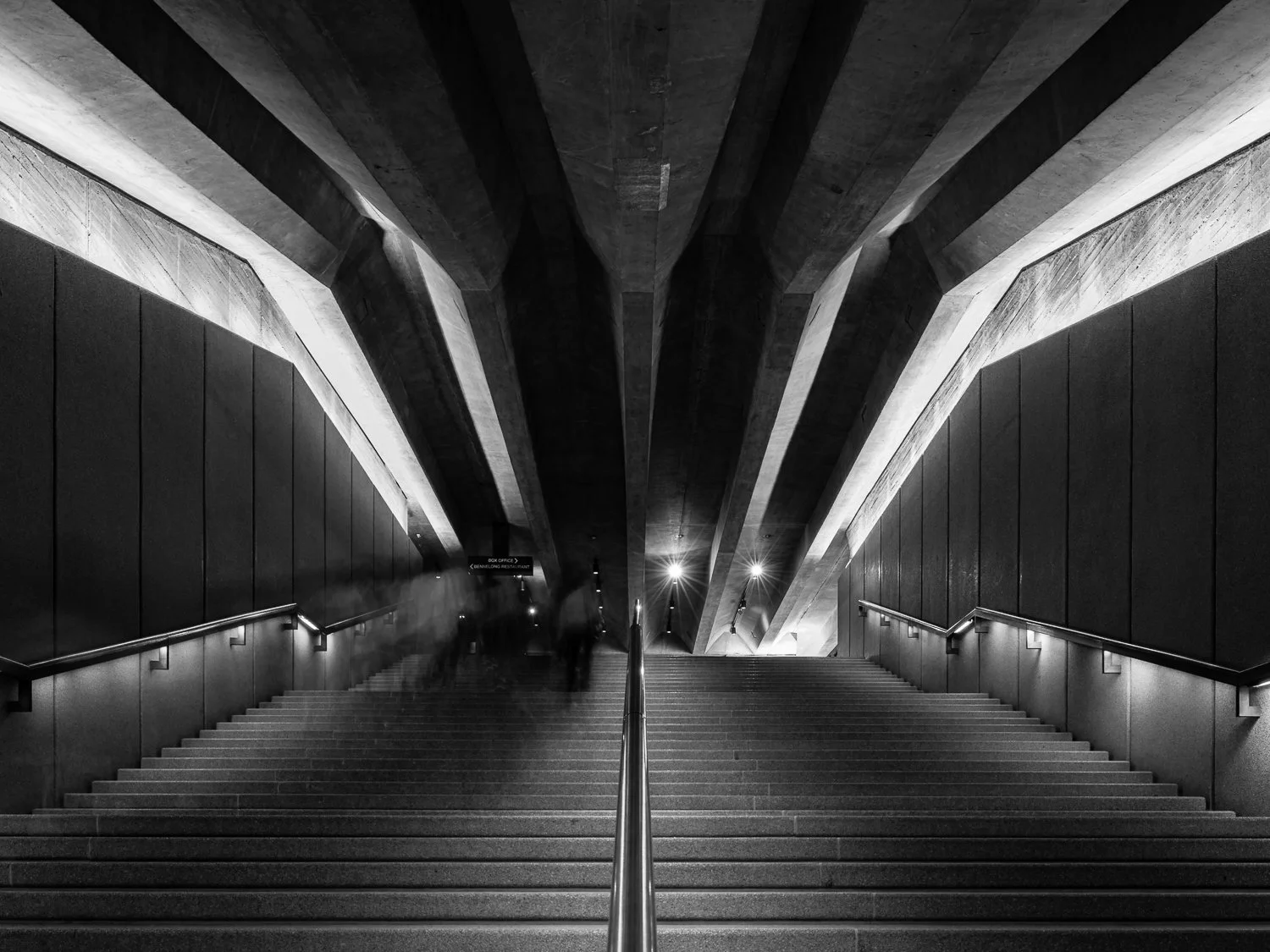Black and white photo of an underground metro station entrance with stairs, walls, and ceiling made of concrete, illuminated by lights on the sides.