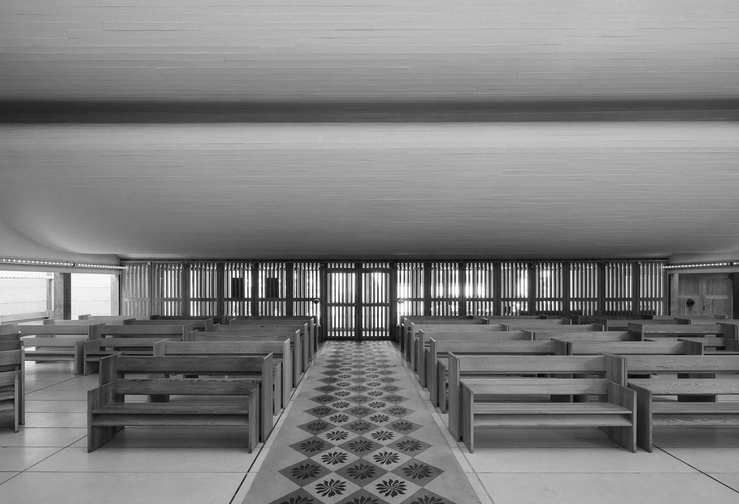 Black and white photo of an empty church pews facing a large entrance with closed wooden doors and vertical wooden slats along the sides, with a patterned tile pathway down the center.