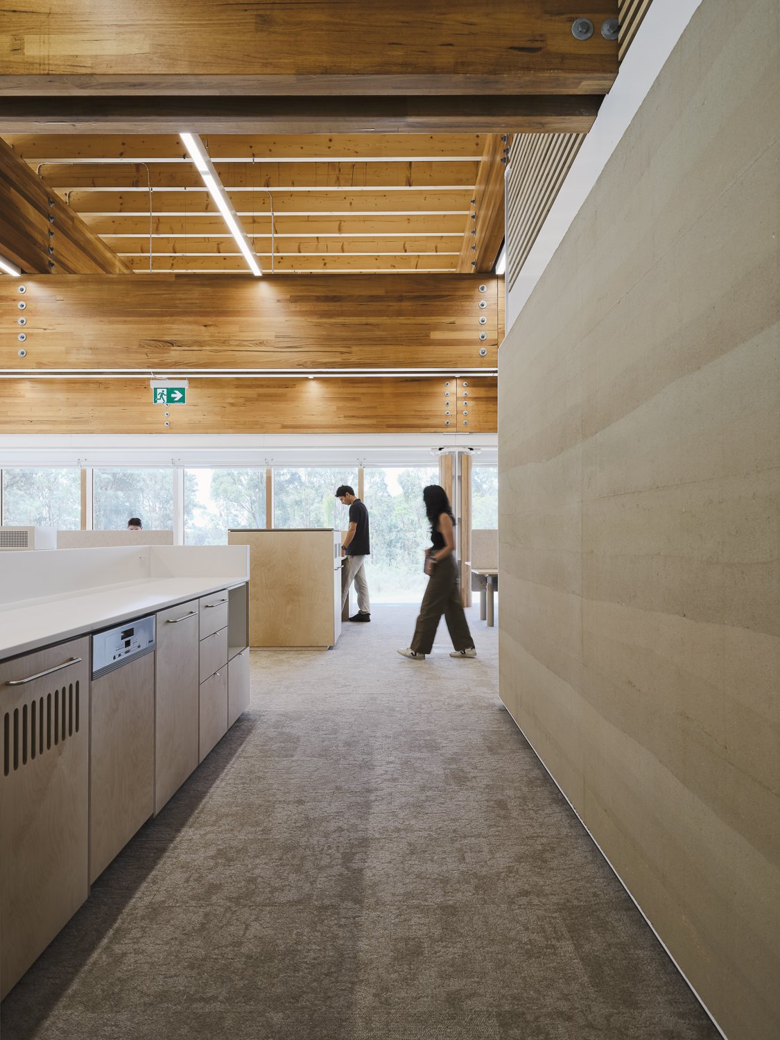 Interior view of a modern office space with wooden beams and an open layout. Two people are walking through the area, which features light wood cabinetry and large windows providing natural light. The ceiling has exposed wooden beams and fluorescent