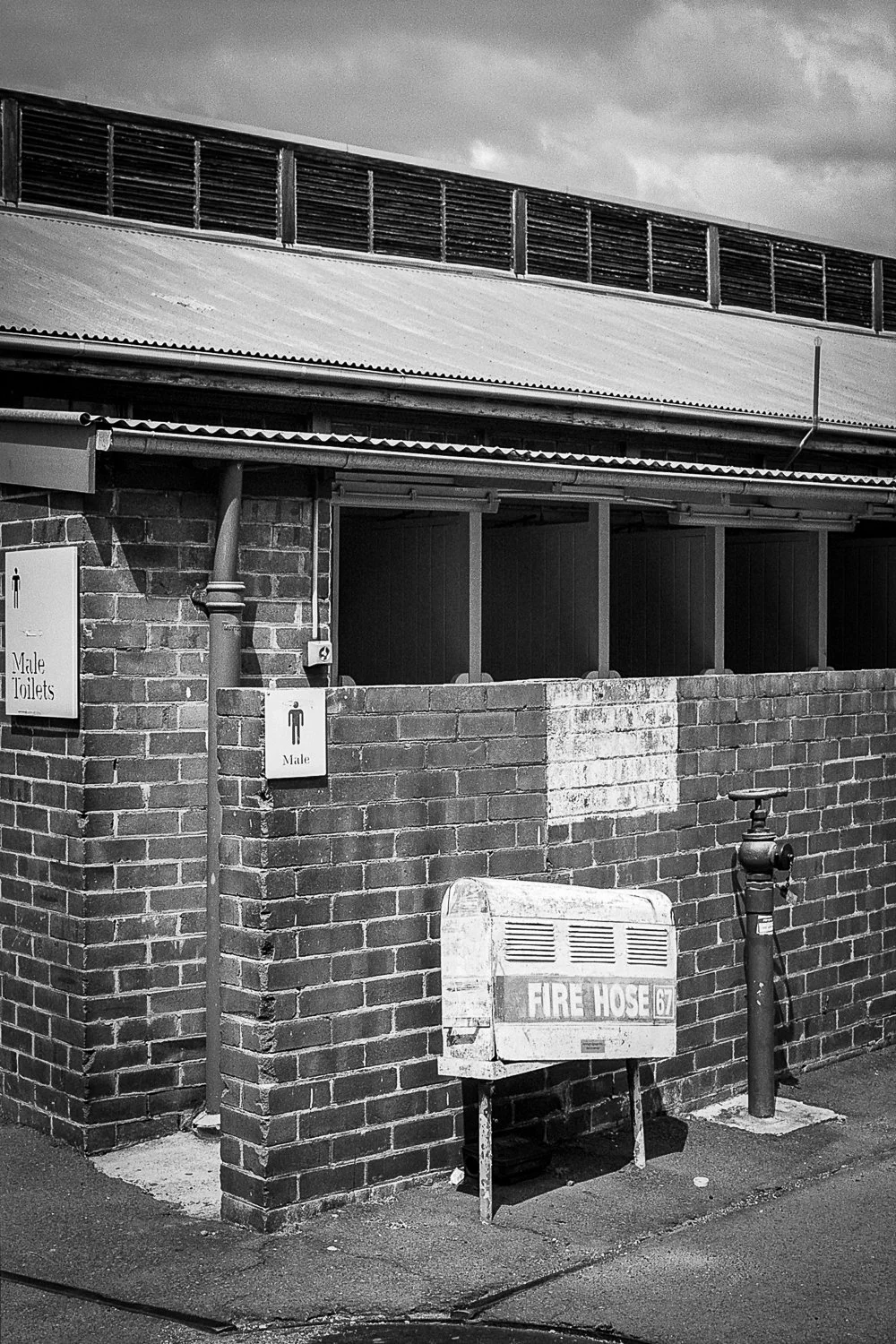 Exterior view of a brick public toilet building with signs indicating male toilets. An old fire hose box and a fire hydrant are outside.