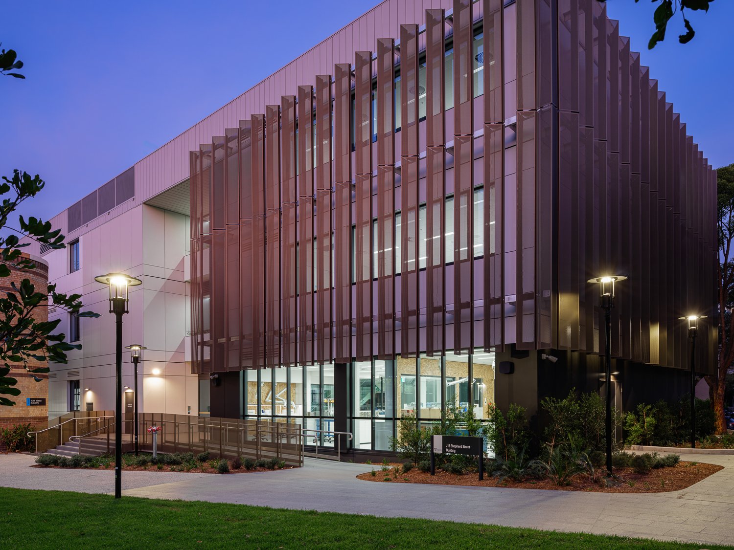 Modern multi-story building with vertical metal panels, large glass windows, illuminated by outdoor lamps at dusk, with landscaped pathway and greenery in the foreground.