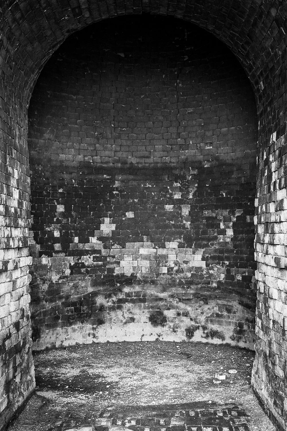 Black and white image of a small, enclosed brick chamber with an arched ceiling, dirt floor, and stained brick walls.