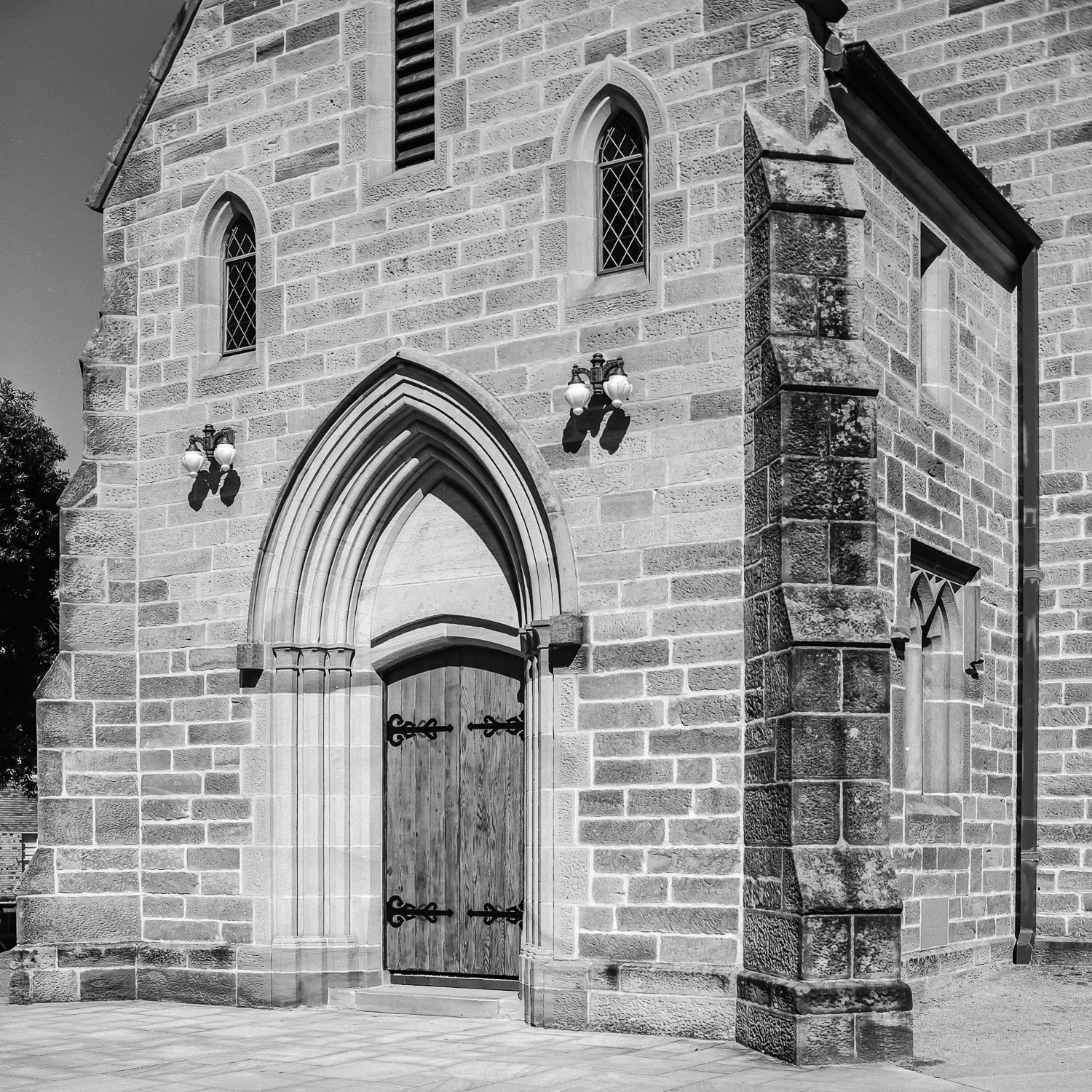 A black and white photo of a stone church with an arched wooden door, small stained glass windows, and exterior wall-mounted lamps.