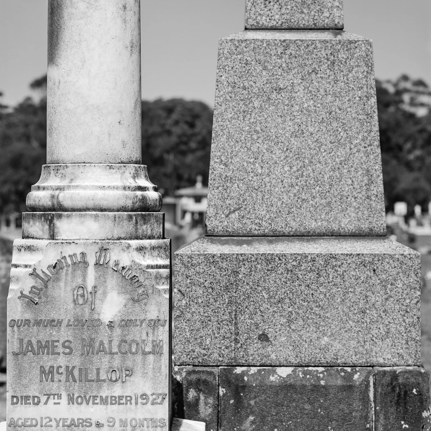Close-up of two sections of a grave monument in a cemetery, one with engraved text and the other plain stone, in black and white.